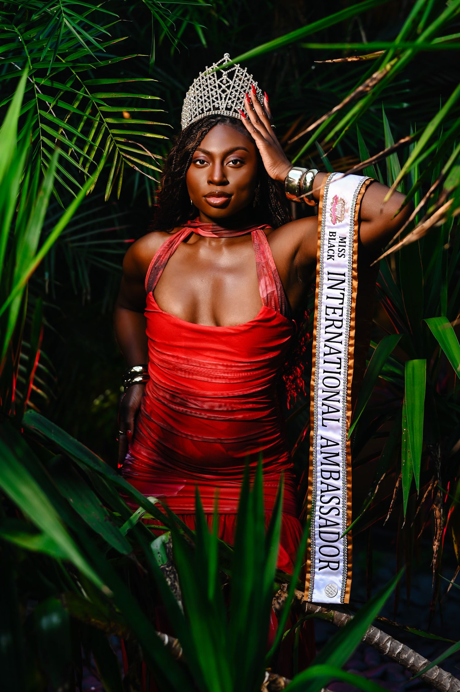Beauty pageant winner in a red gown poses among tropical palm fronds while wearing a crown and sash.