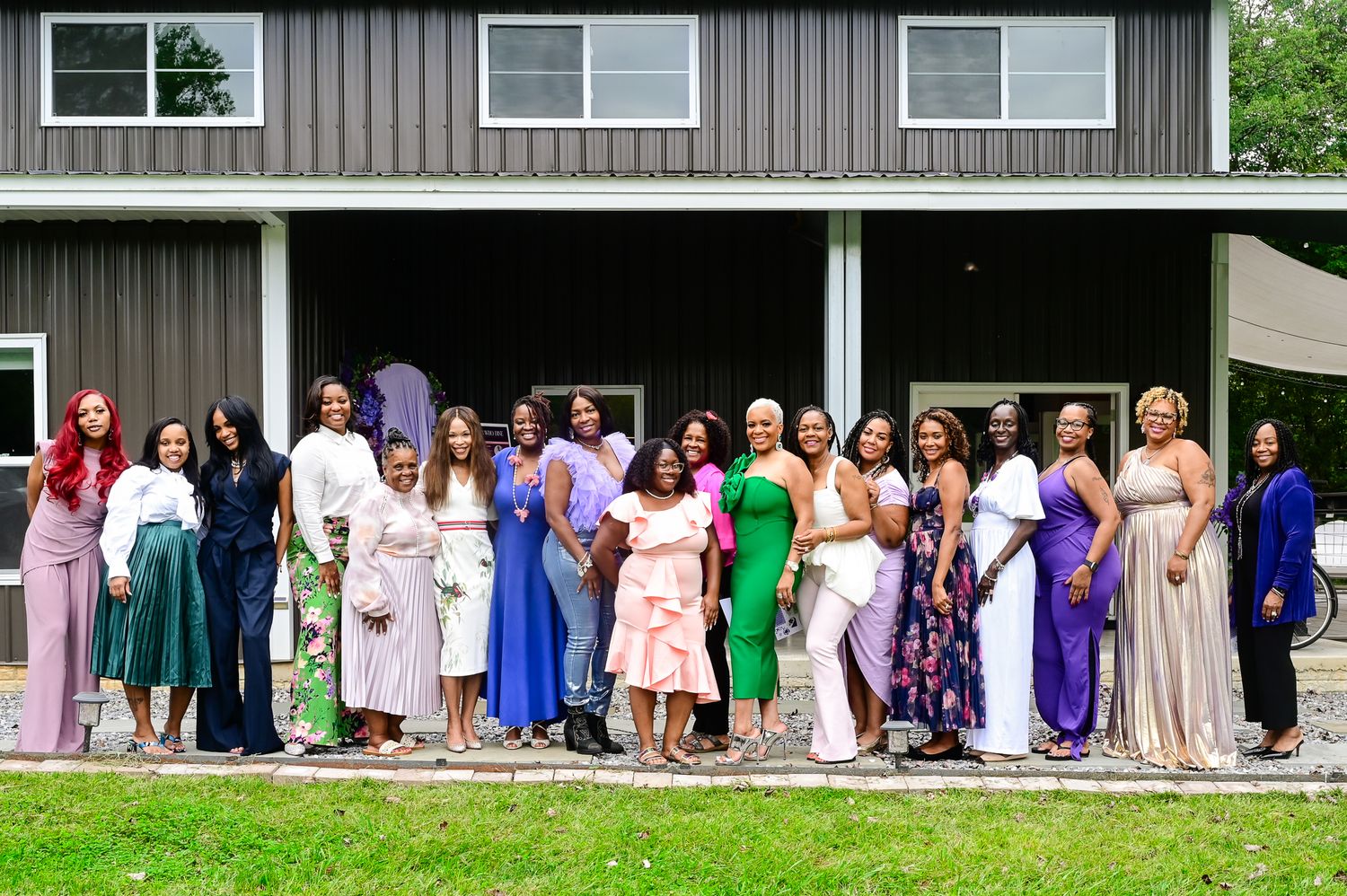 Group of elegantly dressed women posing together outside a gray farmhouse for a special celebration event.