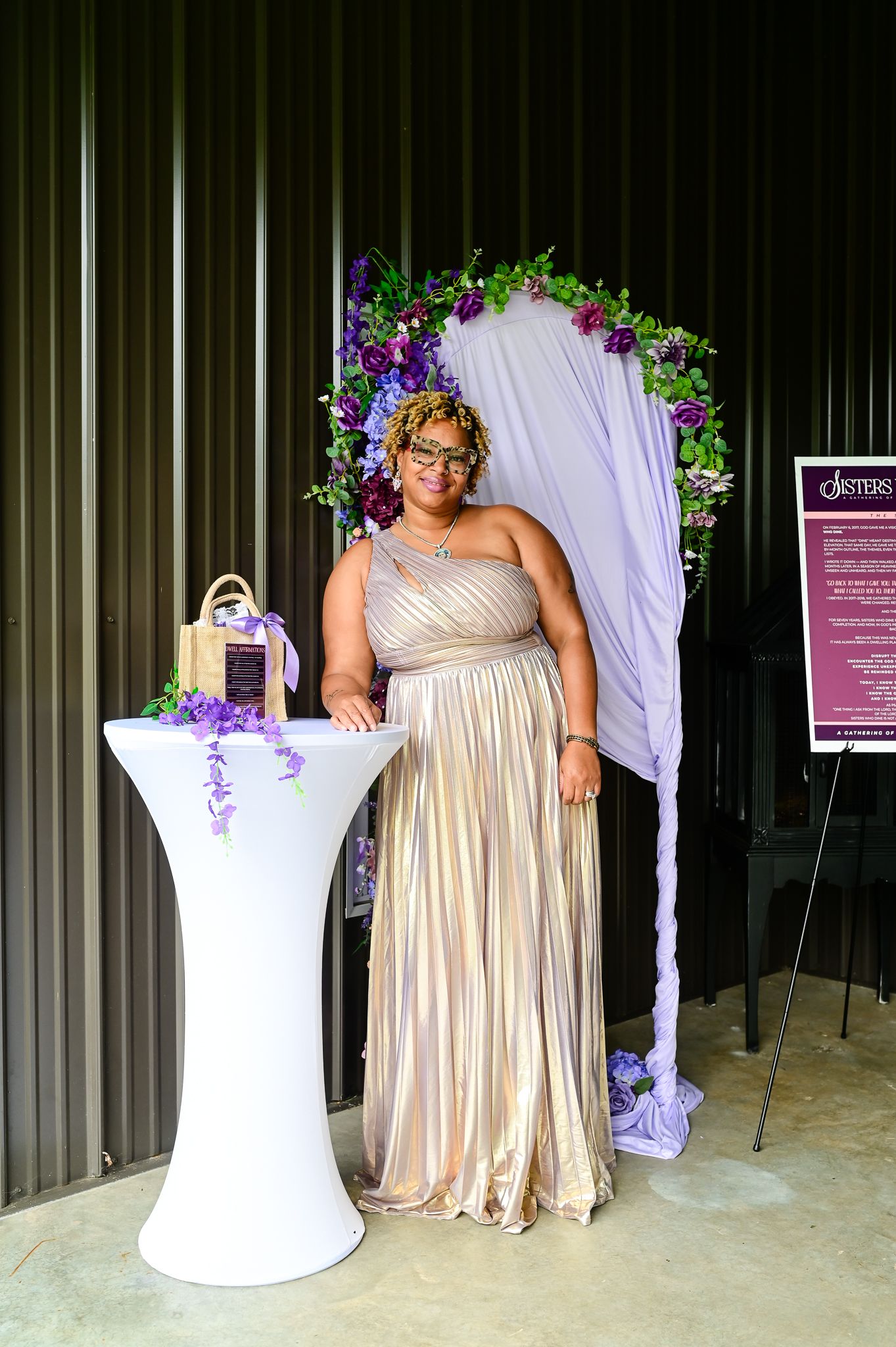 Woman in gold gown poses by floral arch with purple flowers at elegant event.