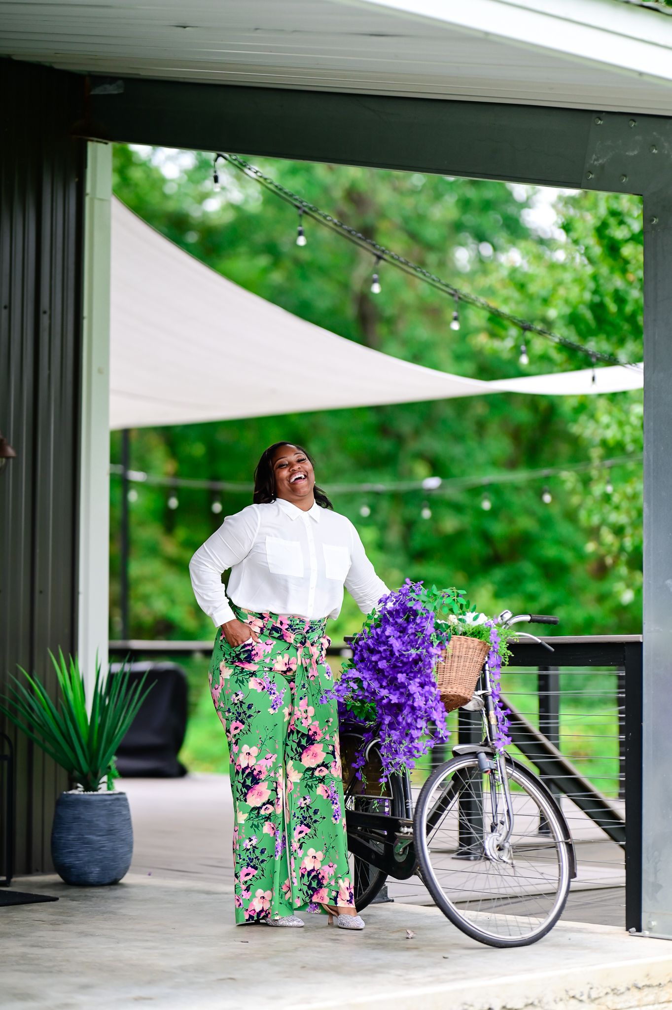 Woman in floral pants and white shirt laughing beside a bicycle with purple flowers on a patio.