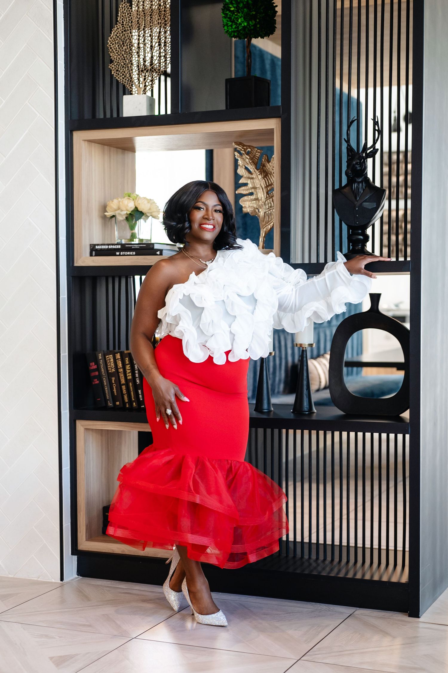 Woman in red mermaid skirt and white ruffled top posing elegantly by a stylish black bookshelf.