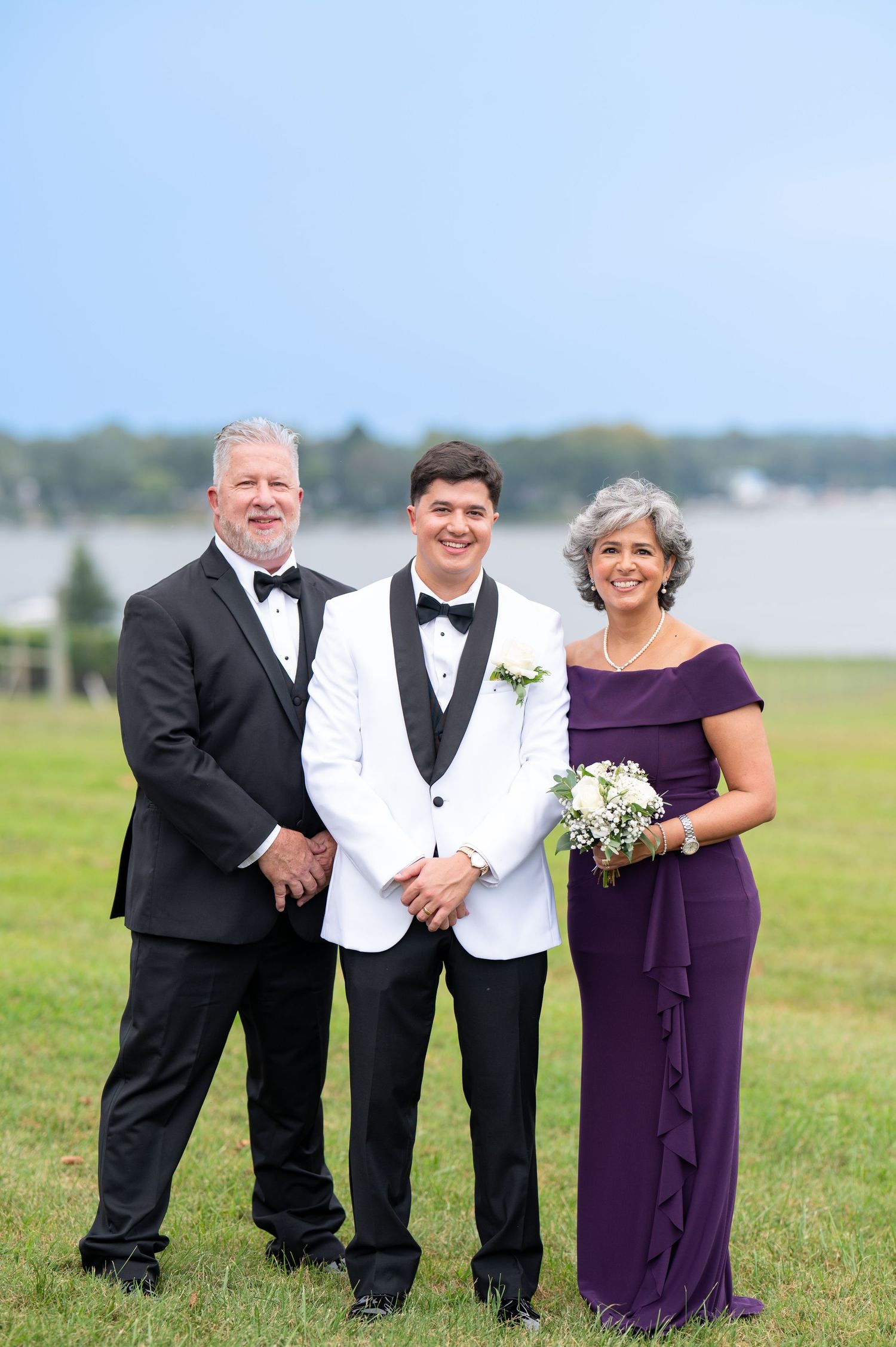 Three people in formal attire pose for a photo on a grassy lawn with a waterfront view in the background.