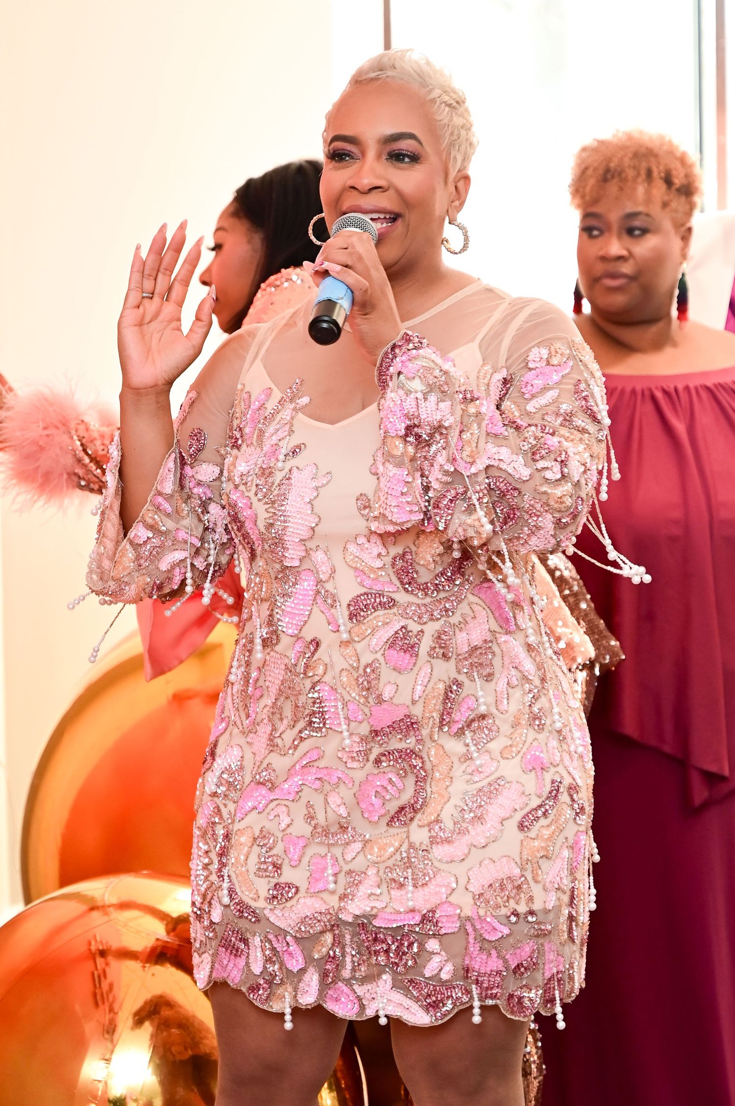 A performer in a sparkly pink sequined dress holds a microphone while singing on stage with backup singers.