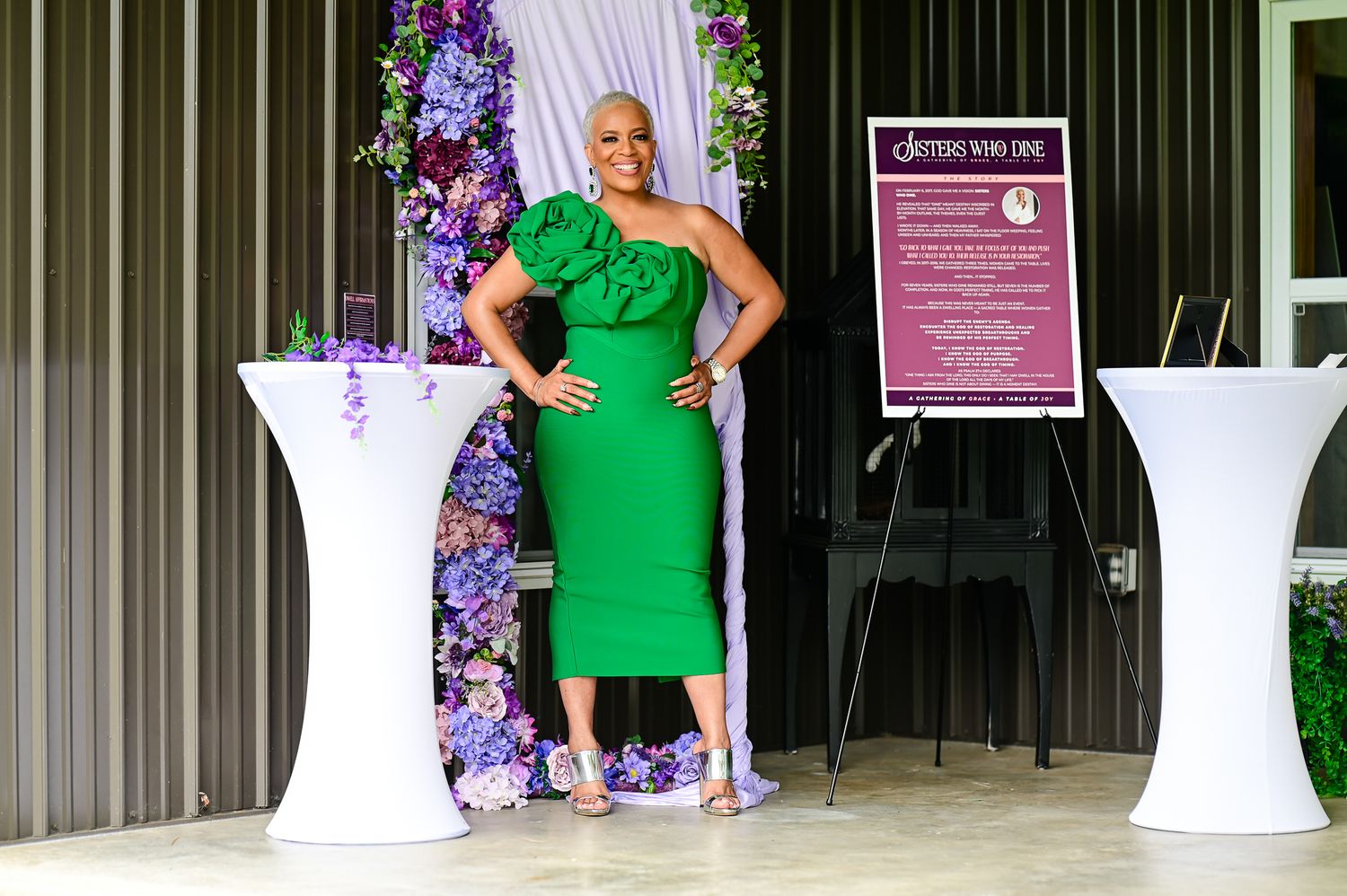 Woman in green one-shoulder dress posing at elegant floral event venue with purple flowers and signage.
