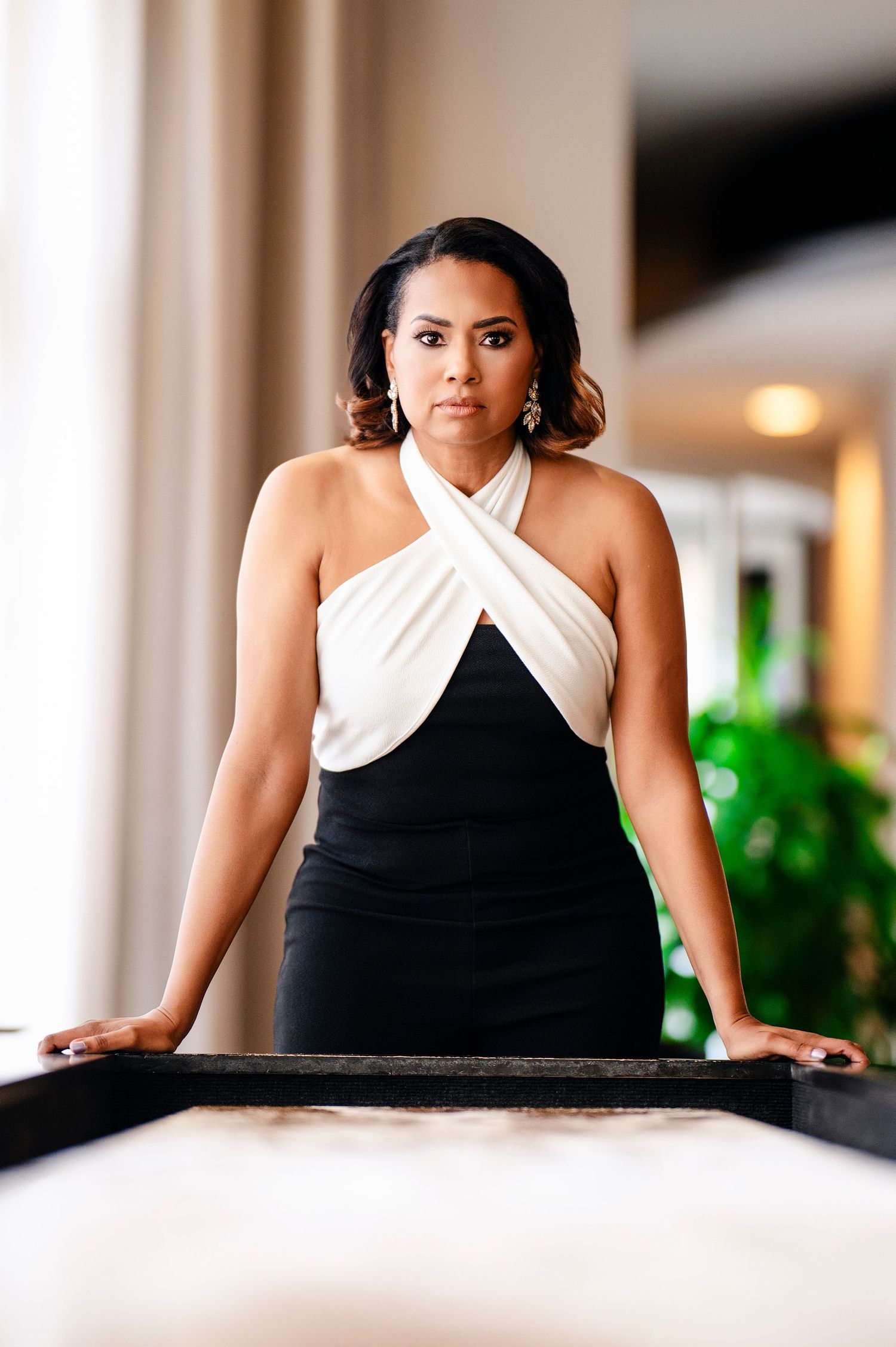 Professional portrait in black and white halter dress standing confidently at a table in an elegant indoor setting.