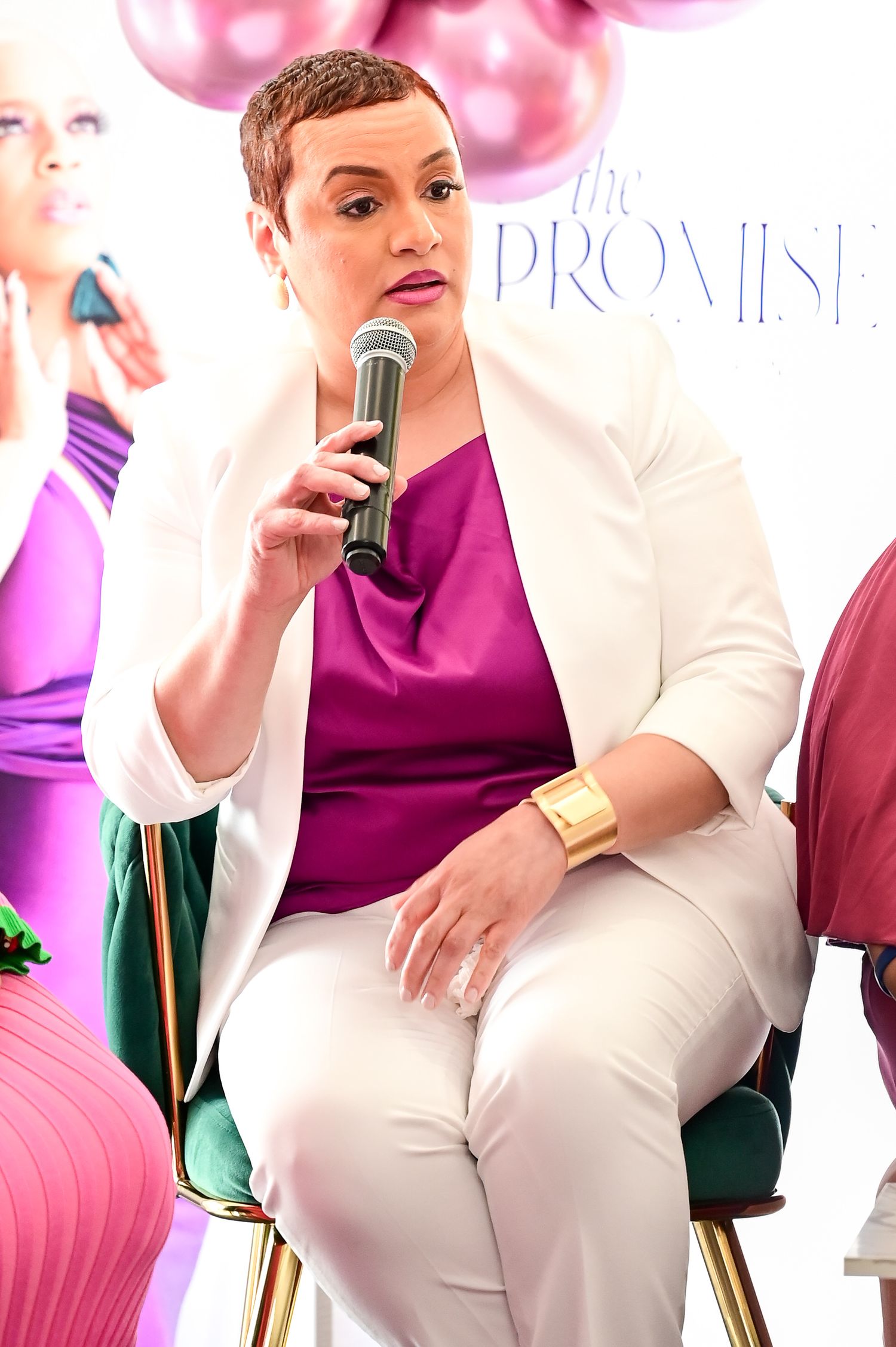 A speaker wearing a white blazer and magenta top sits in a chair with pink decor and balloons in the background.