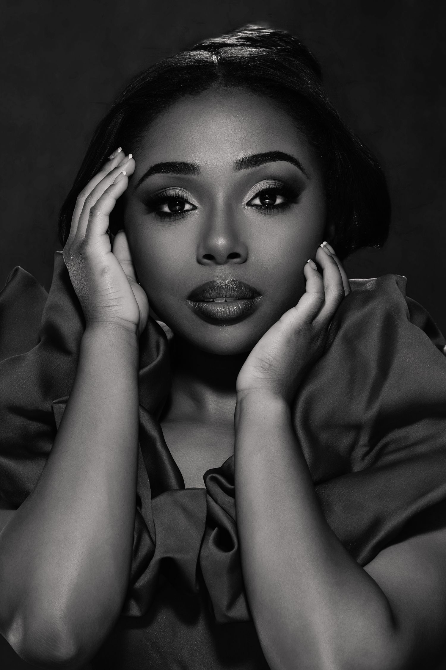 Black and white portrait of a glamorous woman with elegant makeup, hands framing her face, wearing a satin ruffled top.