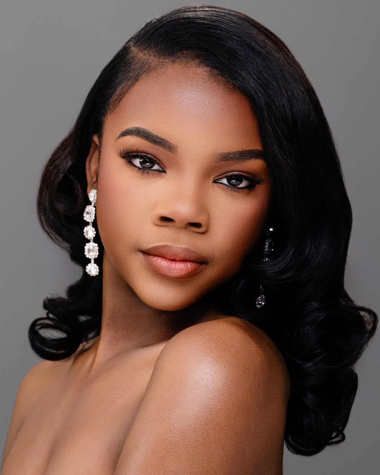 Professional beauty portrait with elegant makeup, flowing black hair, and dangling pearl earrings against a gray backdrop.