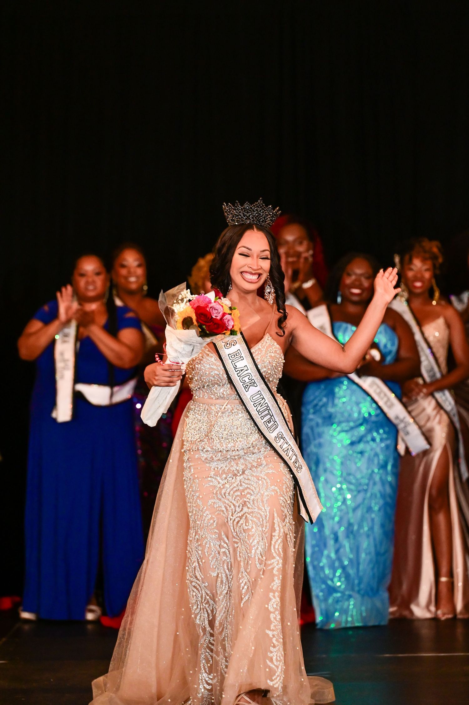 A pageant contestant in a shimmering peach gown waves and smiles on stage while other contestants stand in the background.