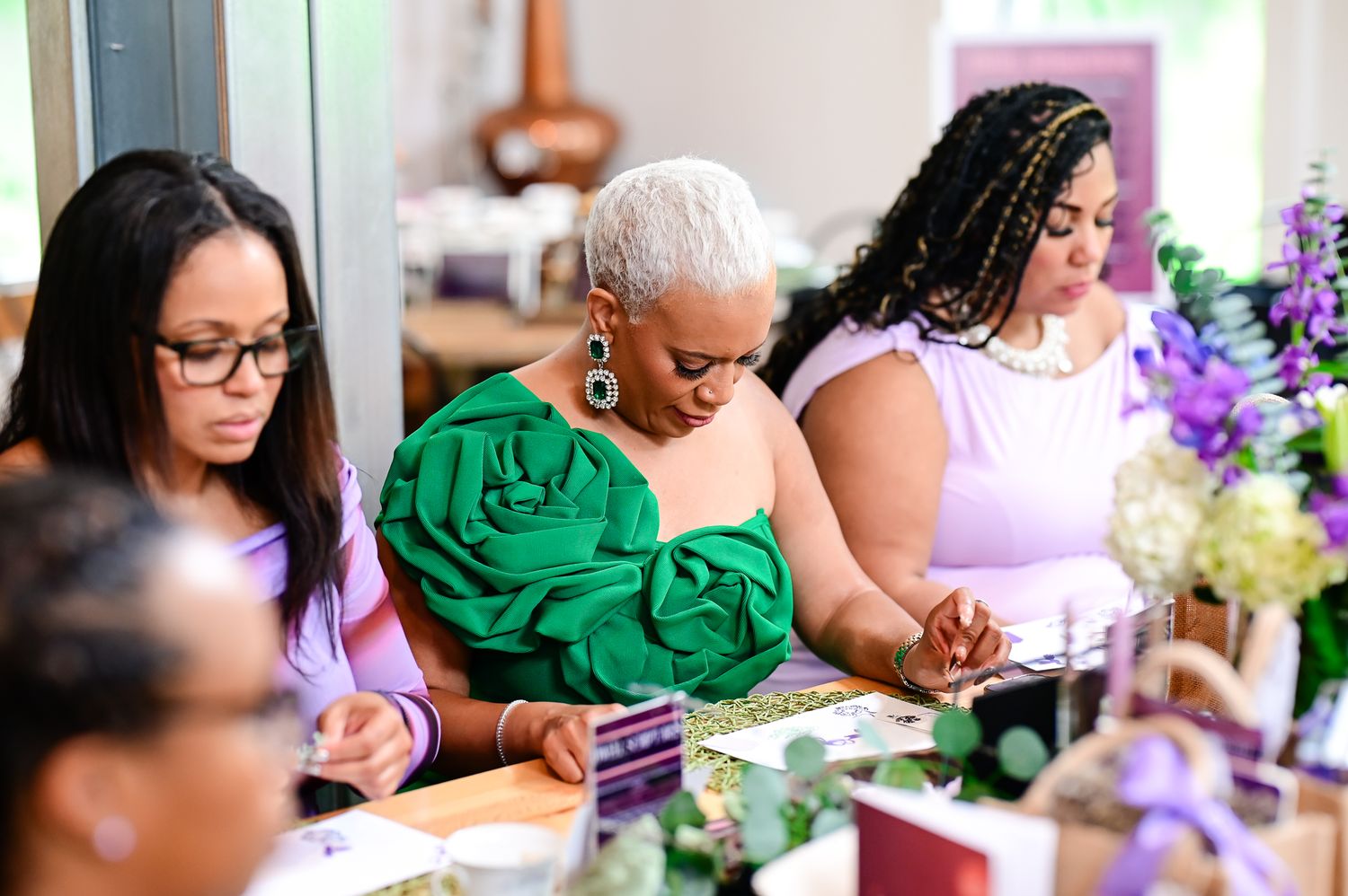 Three women seated at an elegantly decorated table, wearing purple and green outfits at a formal event.