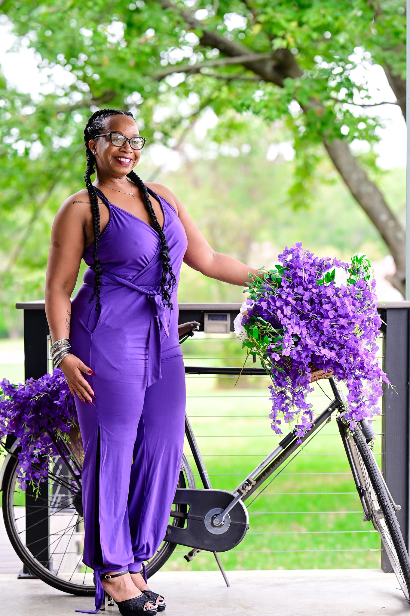 Woman in purple jumpsuit posing with a flower-adorned bicycle outdoors under green trees.