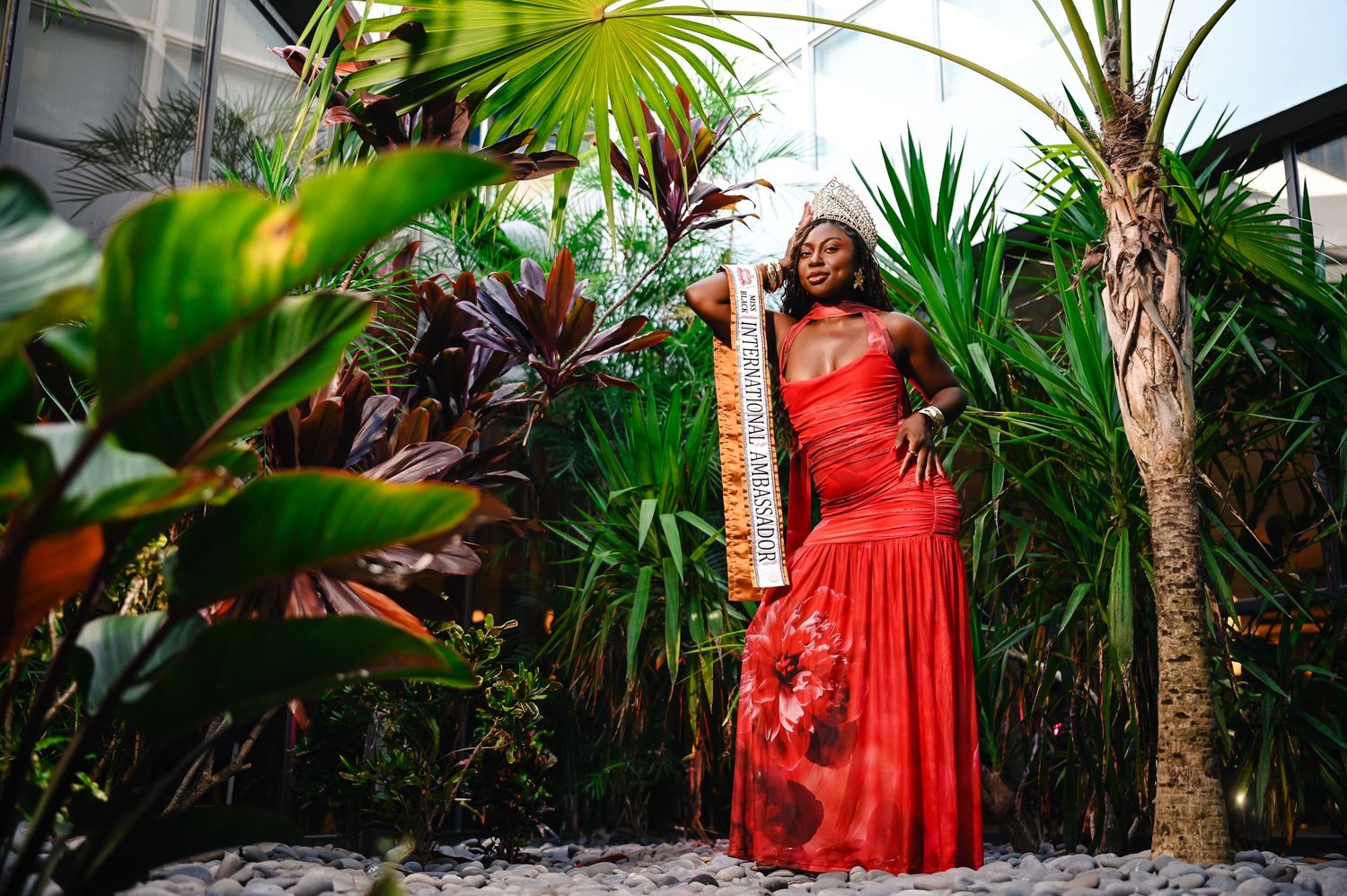 A figure in a flowing red dress poses elegantly among tropical palm trees and lush garden foliage.