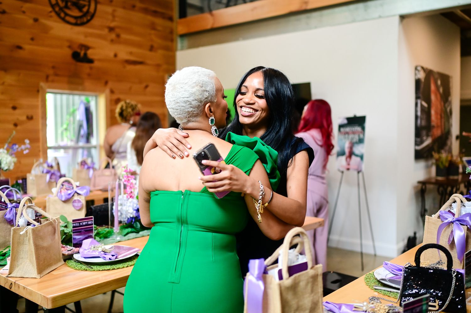 Two women in green dresses share a warm hug at an elegant party with purple gift bags on tables.