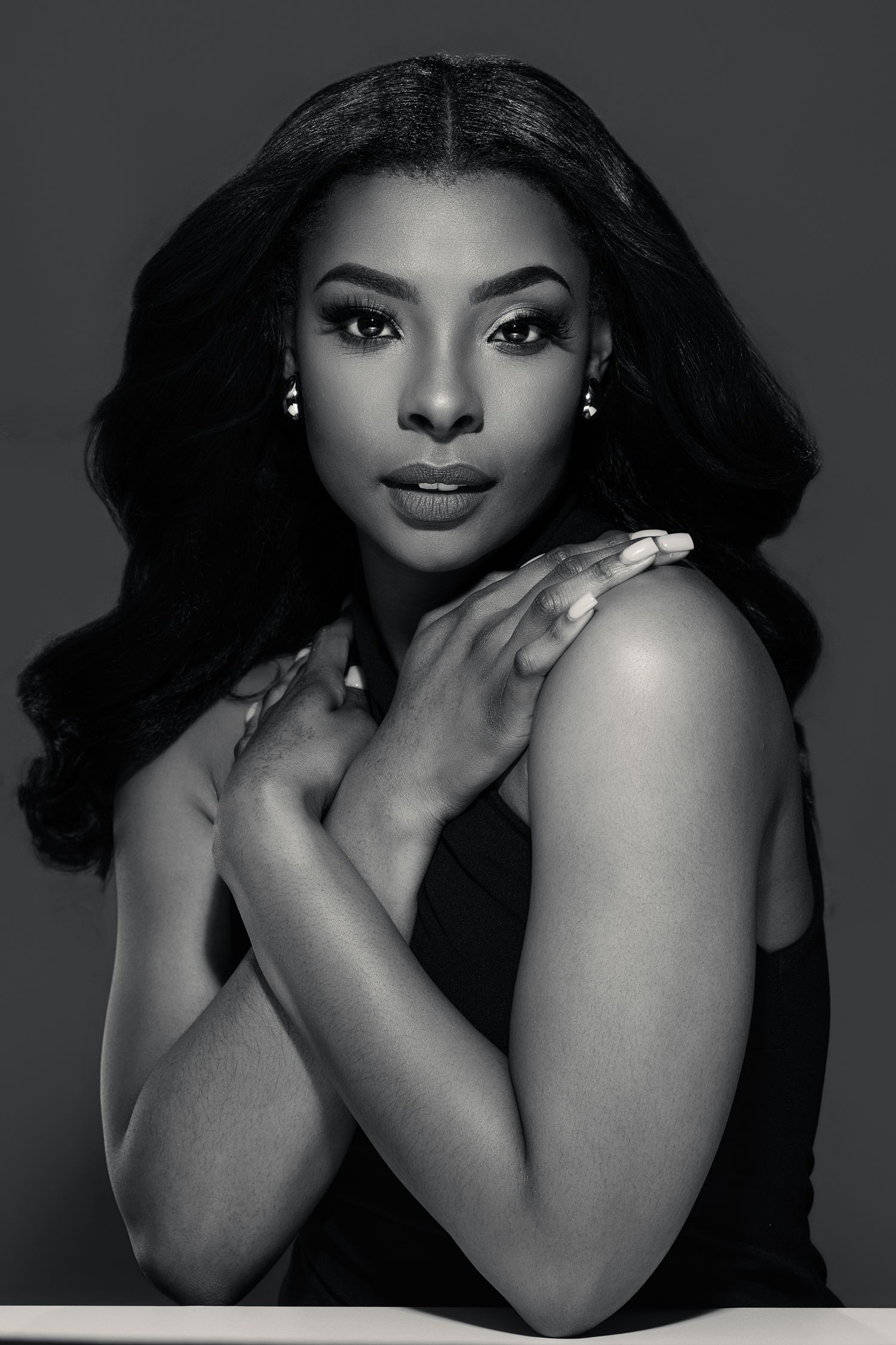 Black and white portrait of a young woman with long wavy hair, crossed arms, wearing drop earrings and a sleeveless top.