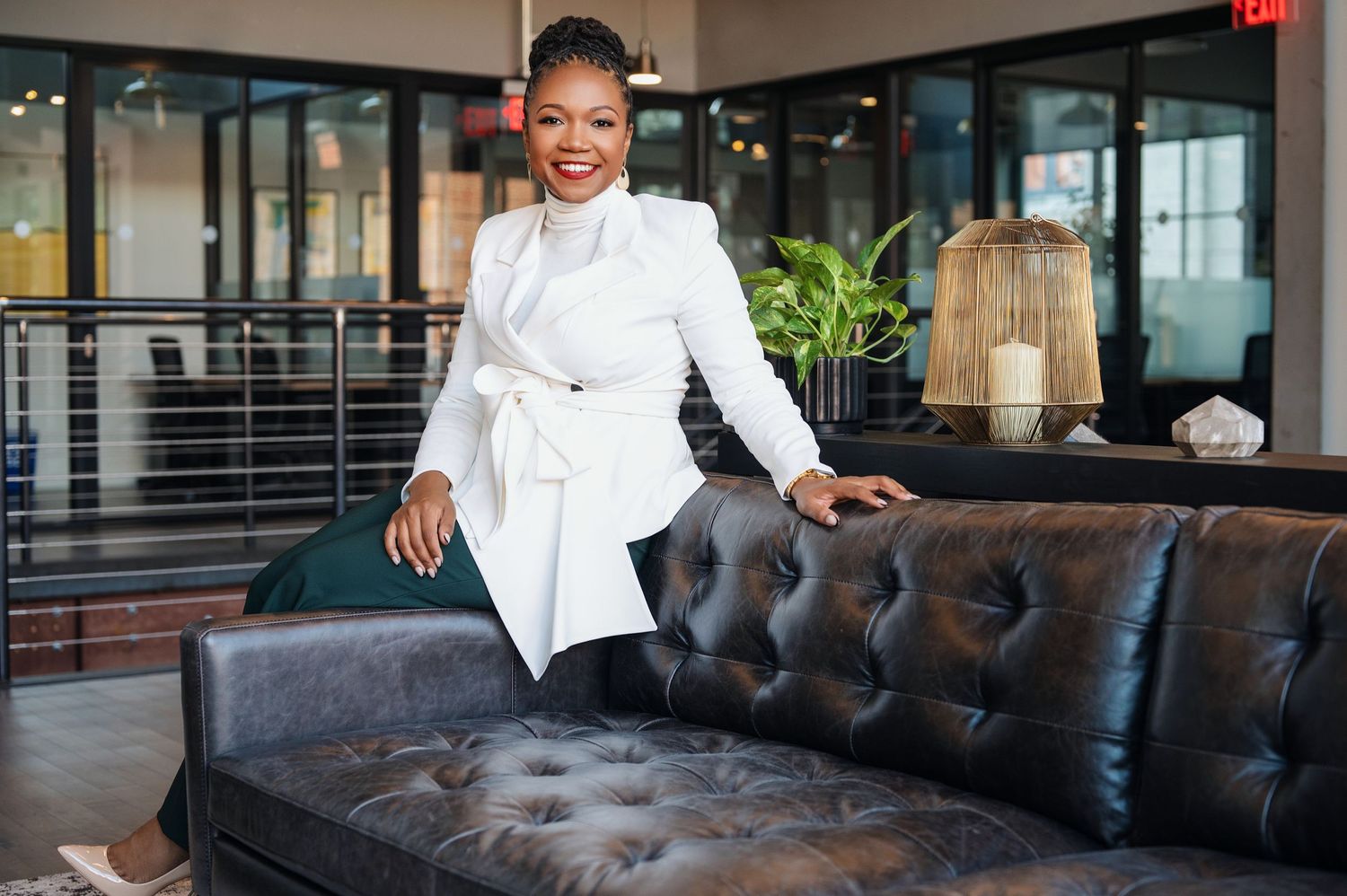 Professional in crisp white blazer smiles while standing next to elegant black leather tufted sofa in modern office setting.