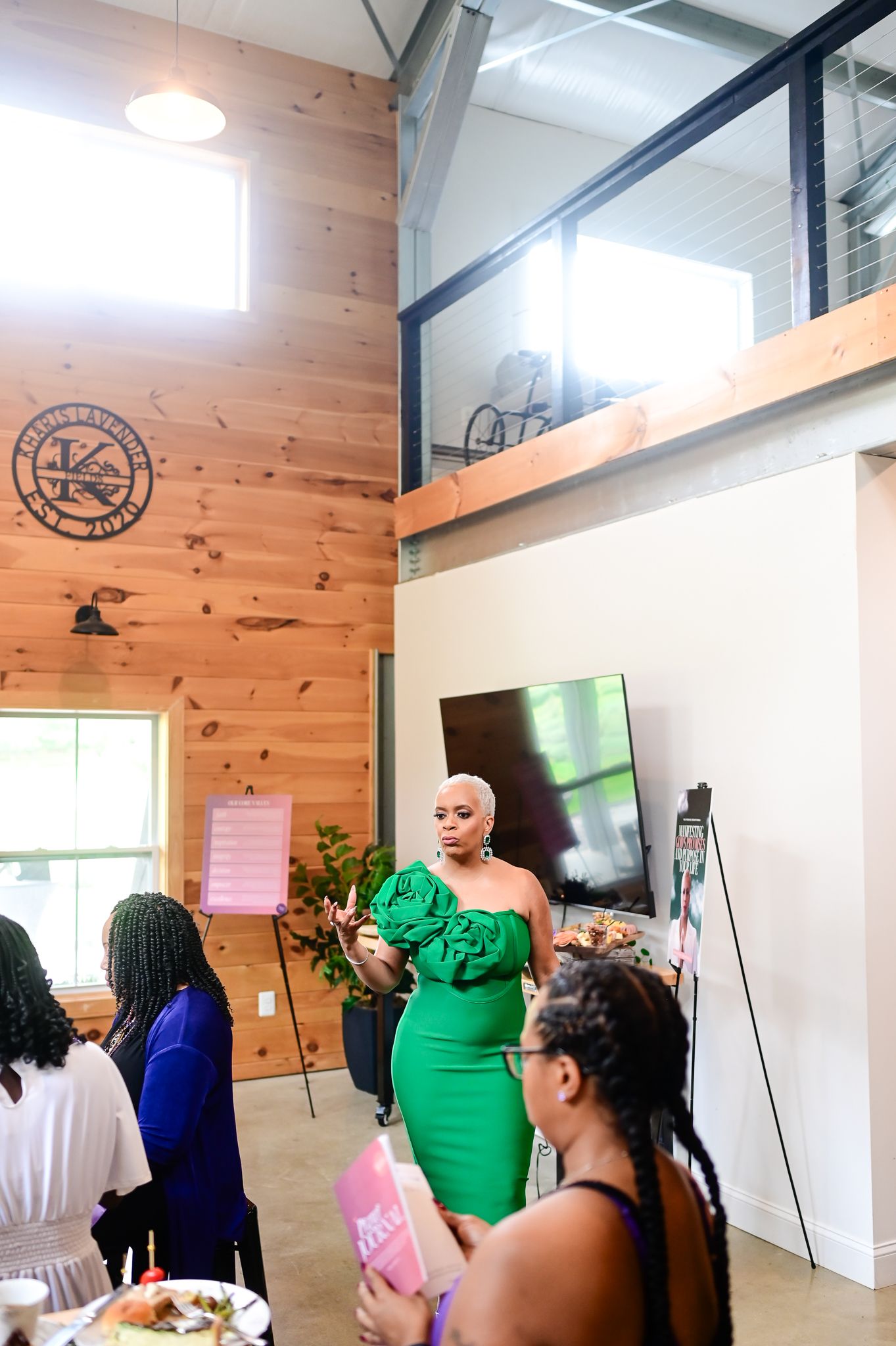 Woman in green off-shoulder dress speaks to seated guests in a rustic wood-paneled event venue.