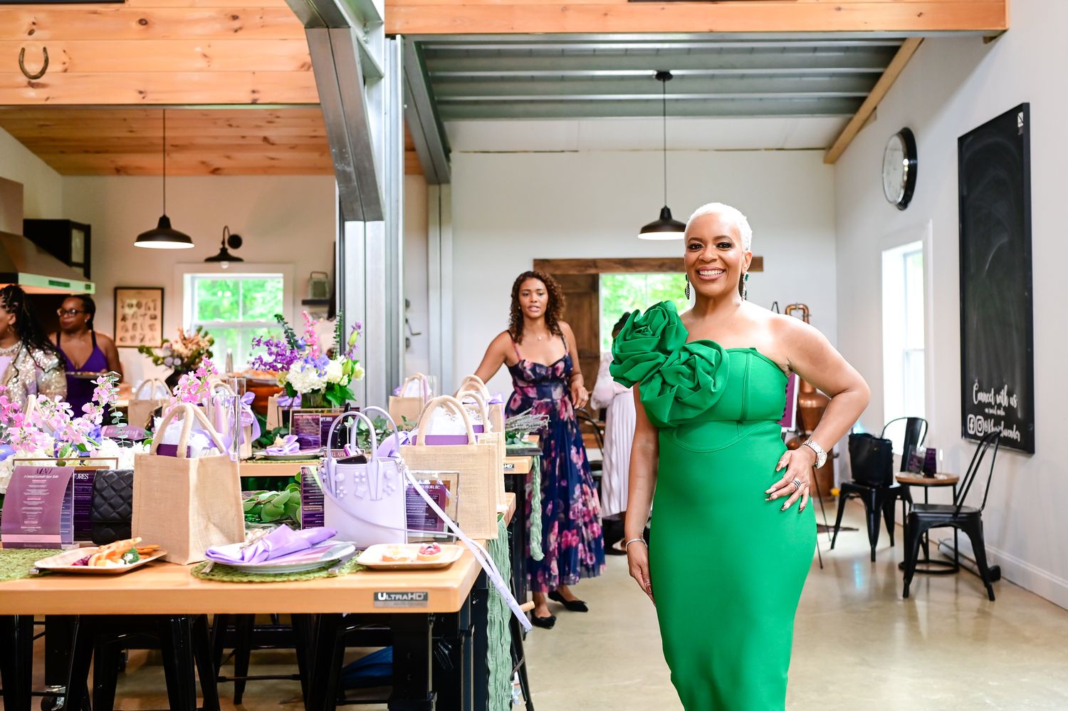 Woman in green dress poses in floral event space with decorated tables and guests in background.