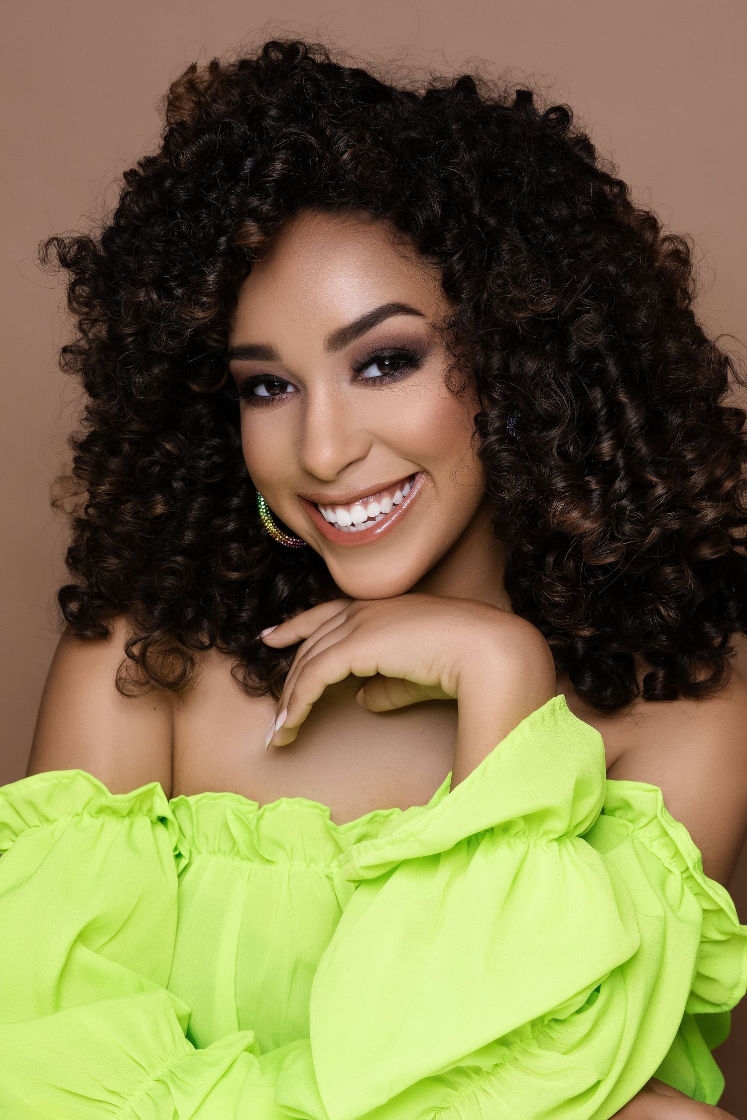 A person wearing a neon yellow ruffled top poses with curly dark hair and a bright smile against a brown background.