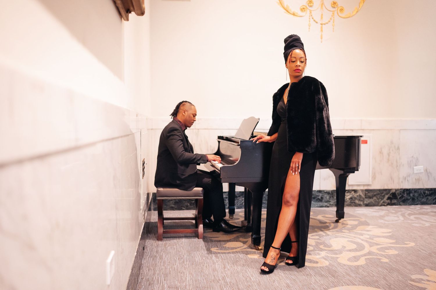 An elegant performer in a black dress gown stands next to a pianist seated at a grand piano in a light-filled room.
