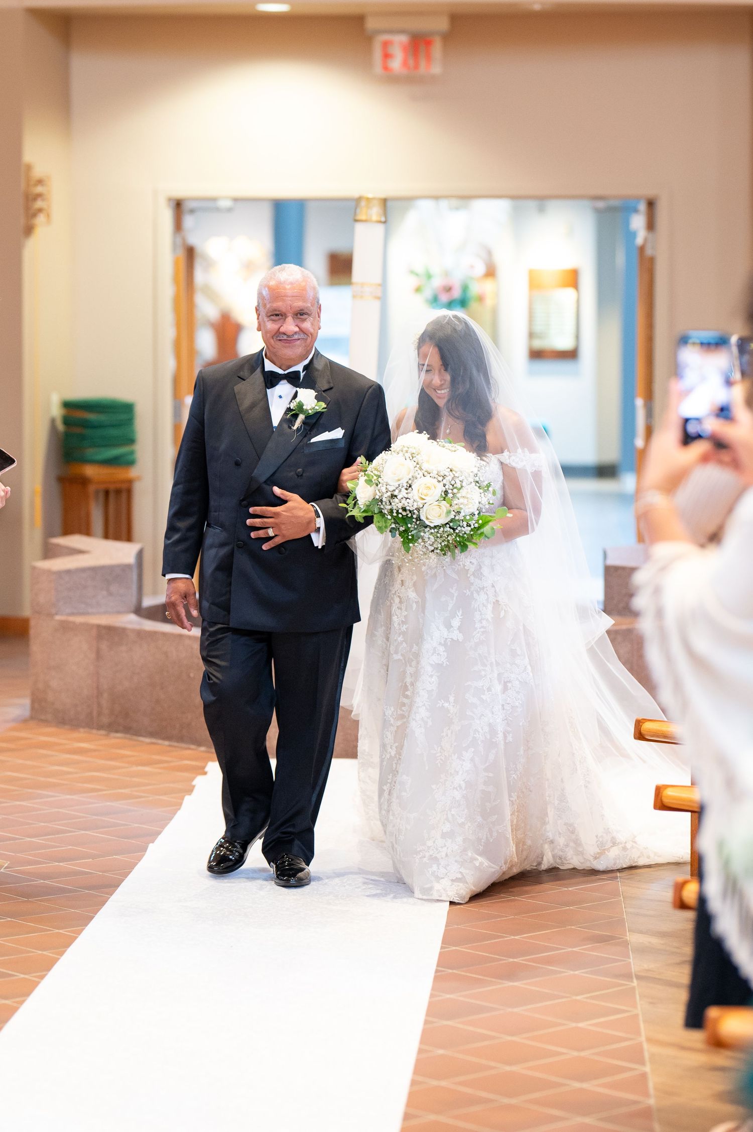 Walking down the white aisle runner at an indoor ceremony venue with a beautiful white bouquet and formal attire.