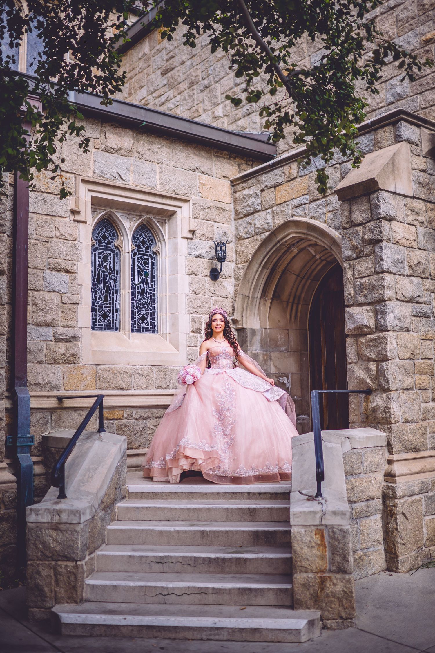 Young woman in pink ball gown poses on stone church steps for quinceañera photos.
