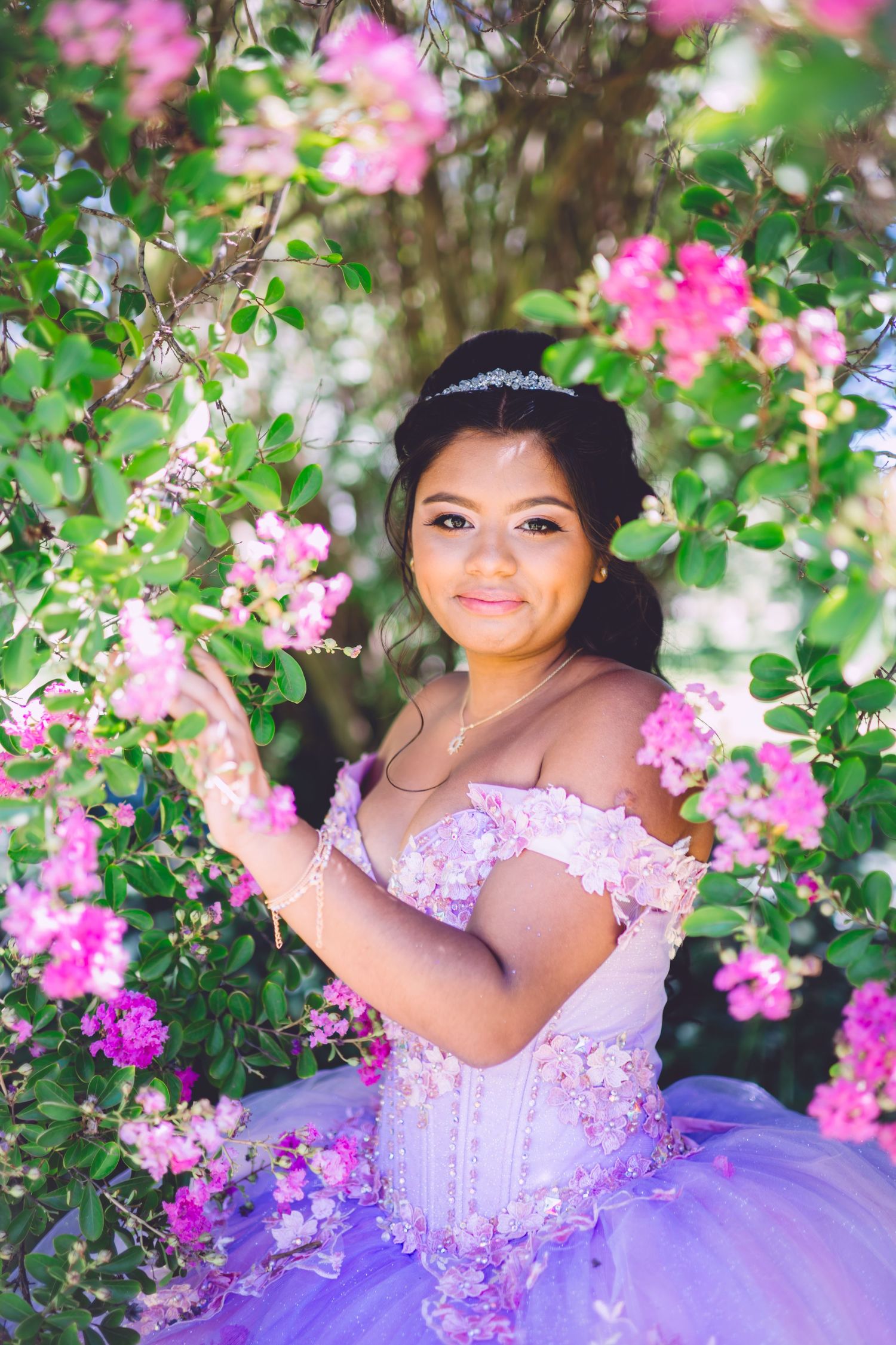 A lavender quinceañera dress sparkles among blooming pink flowers in an outdoor garden setting.