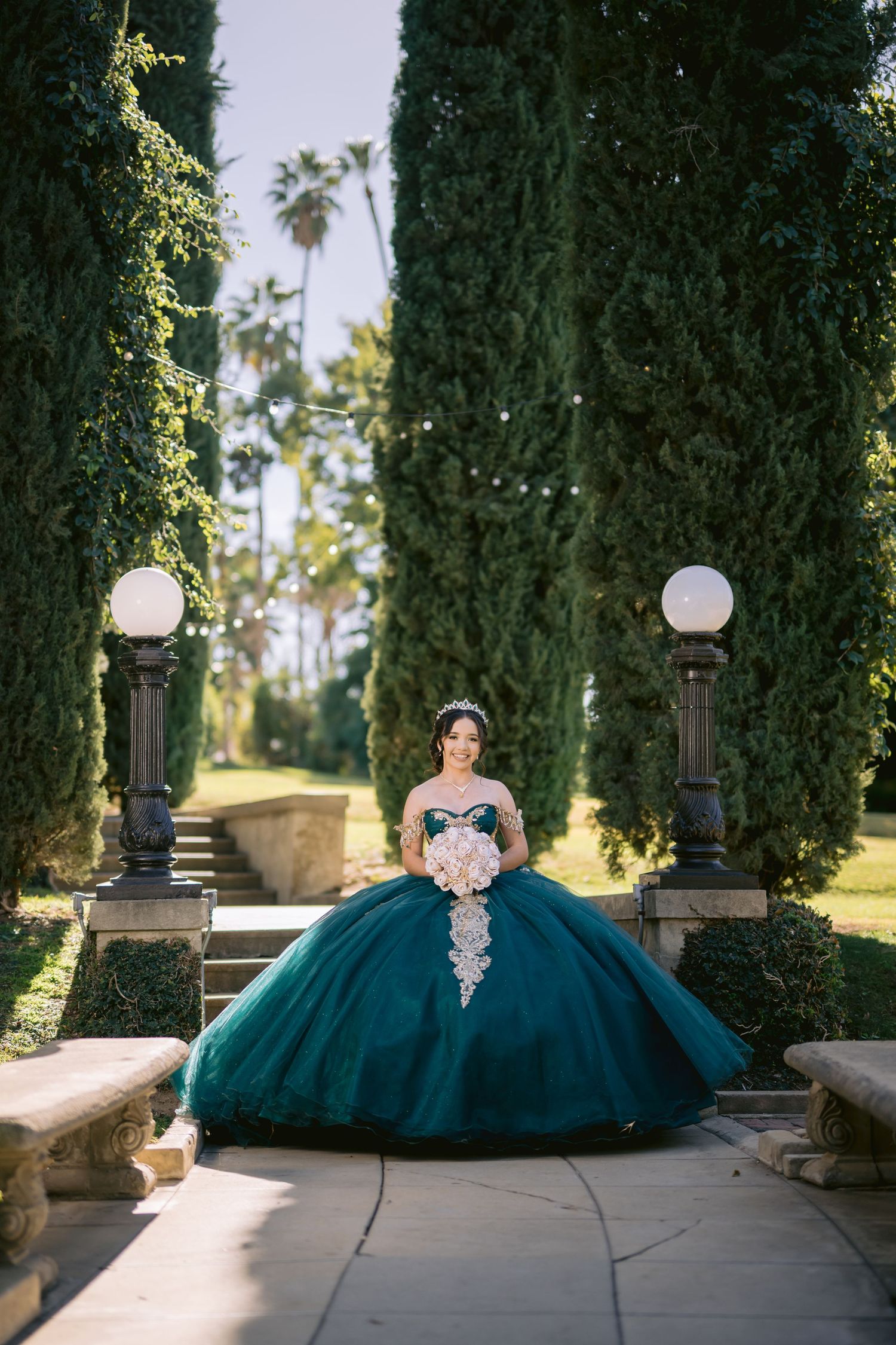 A stunning teal quinceañera ball gown with white lace details photographed in a garden path between cypress trees and lamp posts.