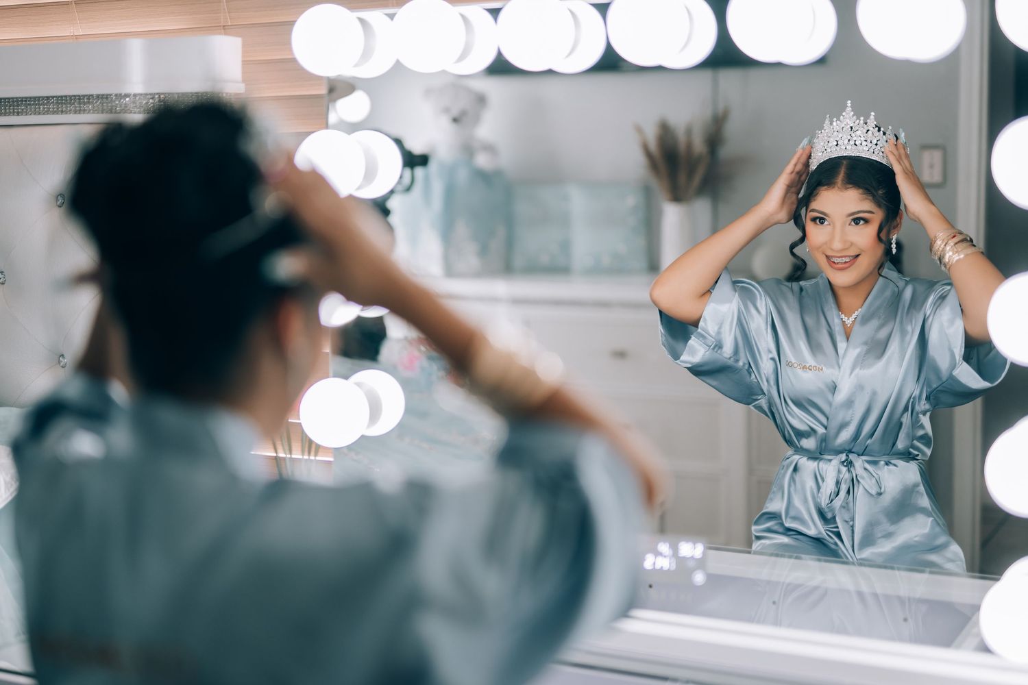 Woman in silver robe placing a tiara crown on her head while looking in a Hollywood vanity mirror.