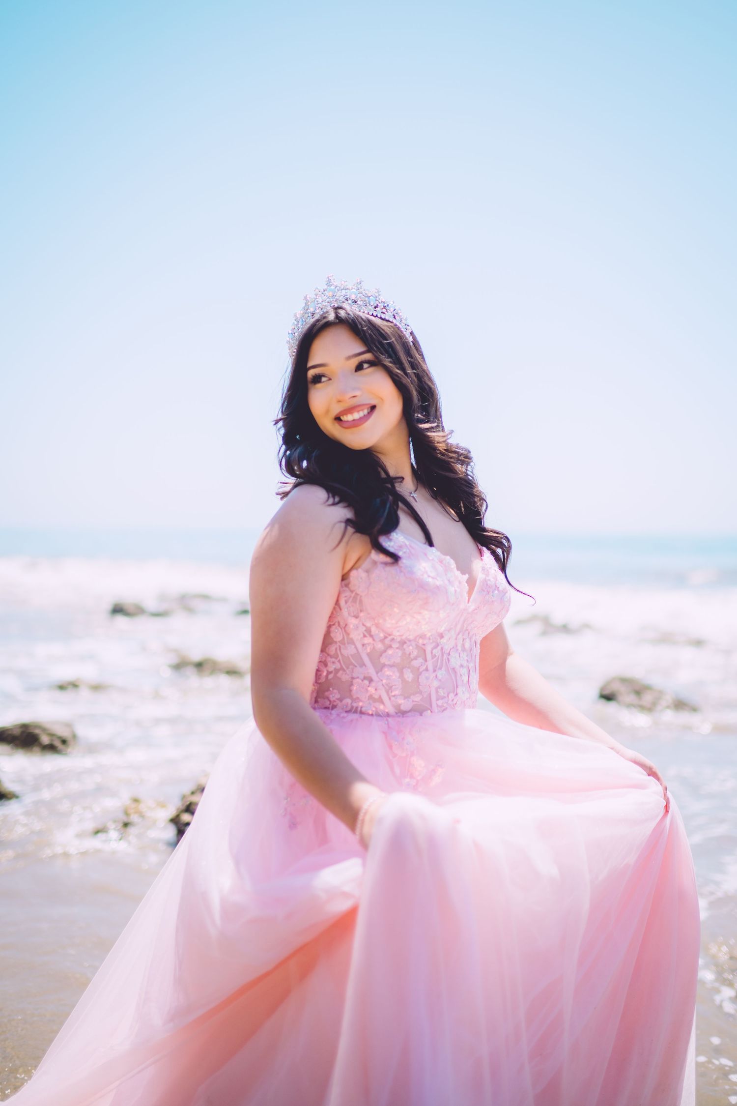 A person in a flowing pink princess gown and tiara poses gracefully on a sunny beach with white sand and blue sky.