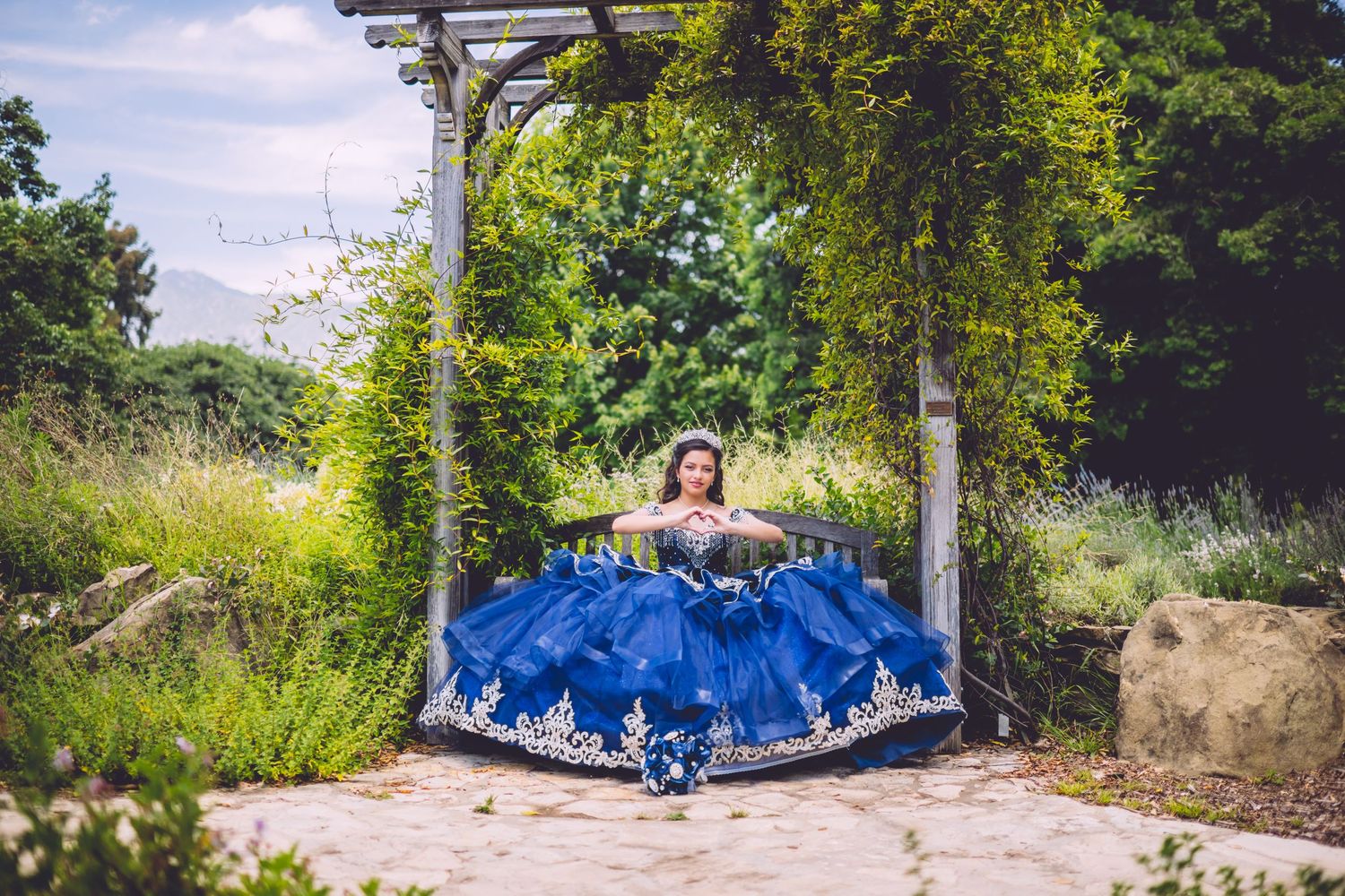 A flowing blue ballgown dress spreads across a garden swing surrounded by lush greenery and flowers.