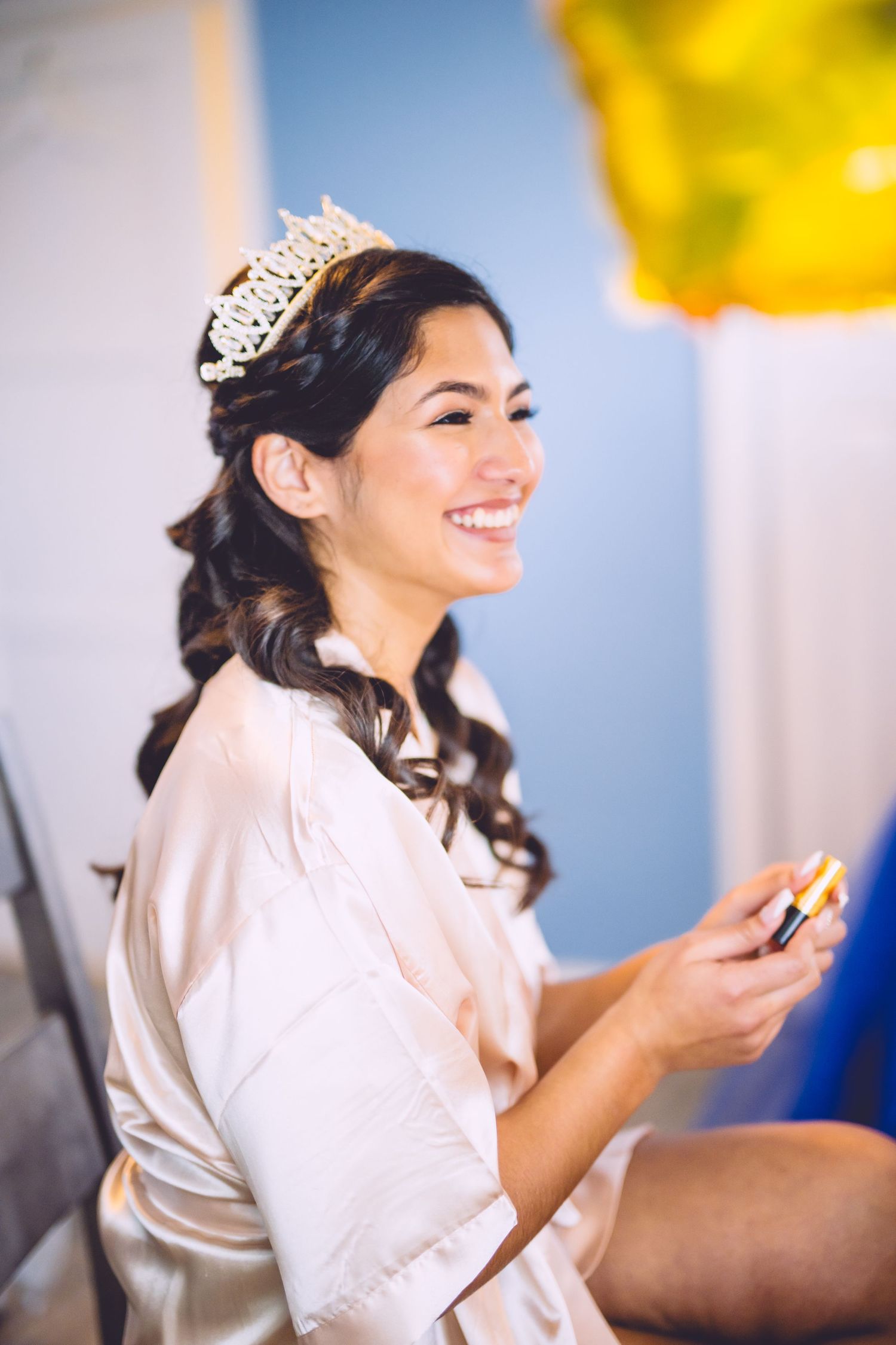 A person wearing a delicate tiara and silk robe smiles while getting ready, with curled hair styled for a special occasion.