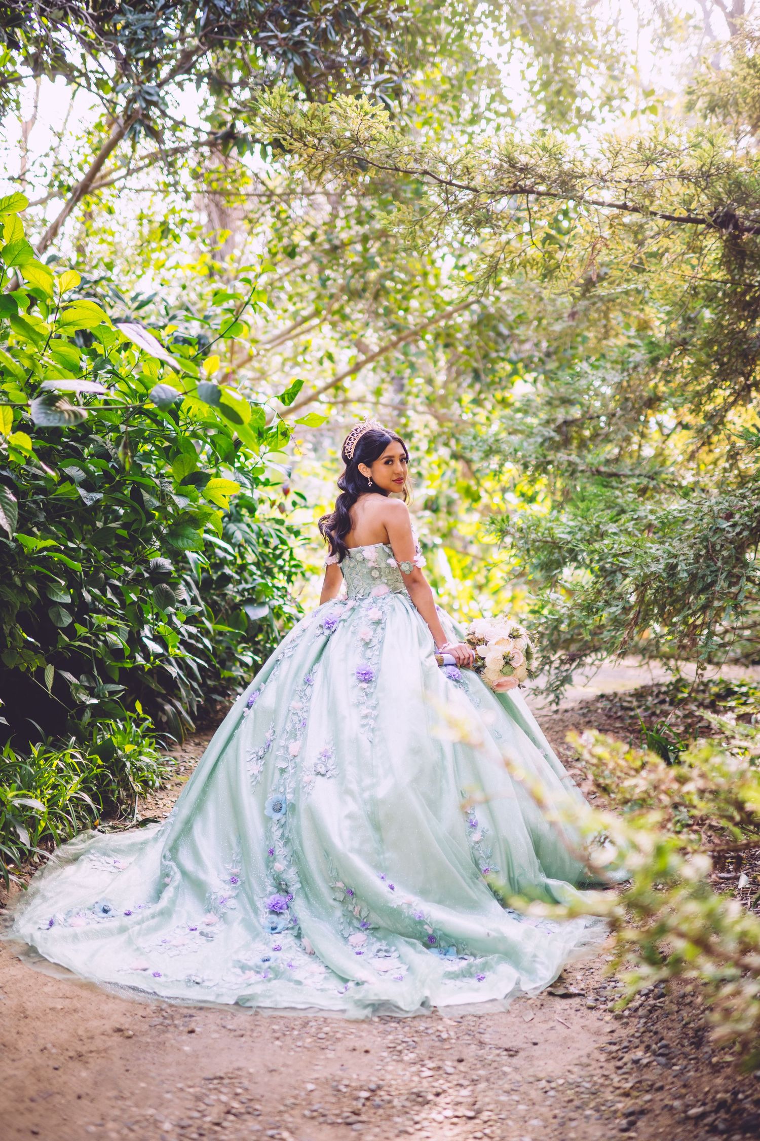 A stunning mint green ball gown with purple floral details catches sunlight on a garden path surrounded by lush foliage.