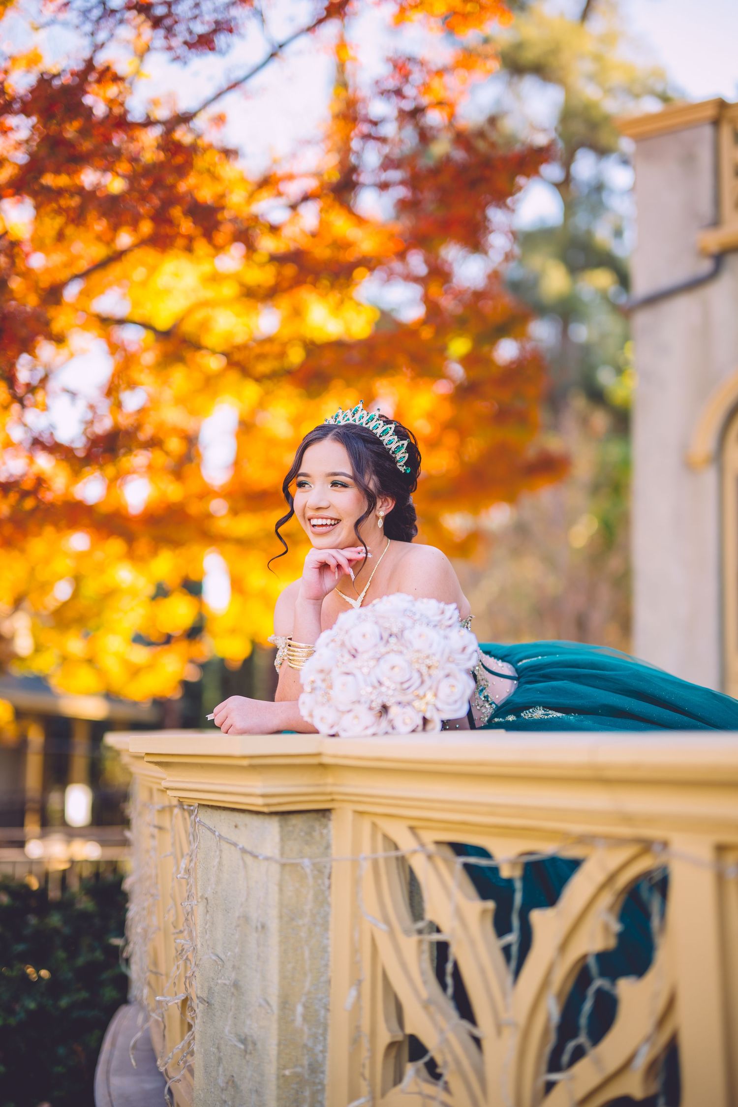 A person in a white lace wedding dress reclines on a cream-colored balcony railing with vibrant autumn foliage in the background.