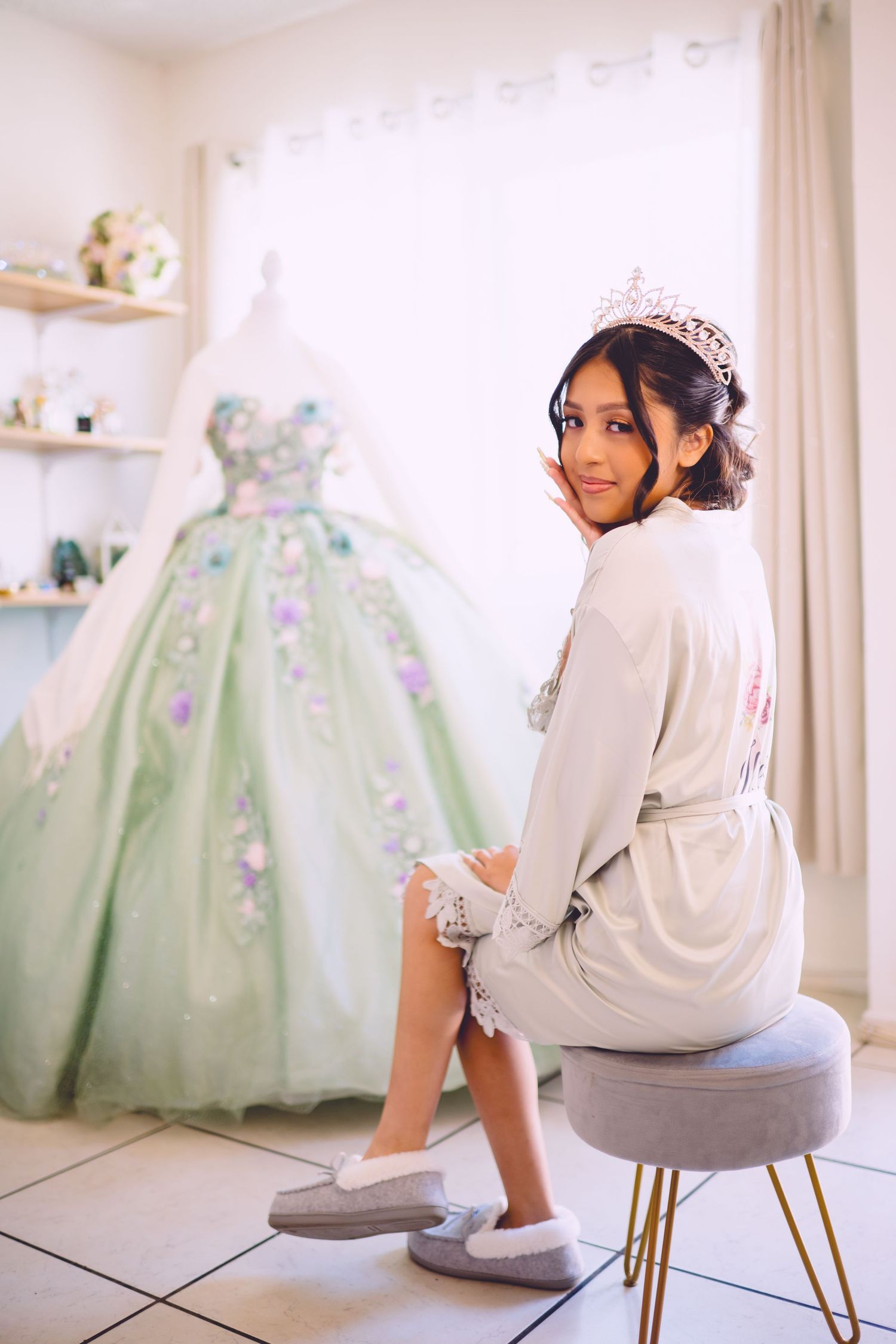 Young woman wearing a tiara and satin robe sits before a floral green quinceañera ball gown on a mannequin.