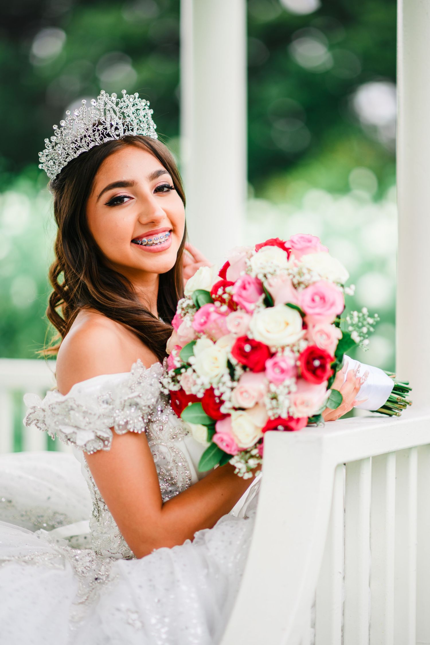 Smiling young woman in white gown and crystal tiara holds colorful bouquet of pink and red roses.