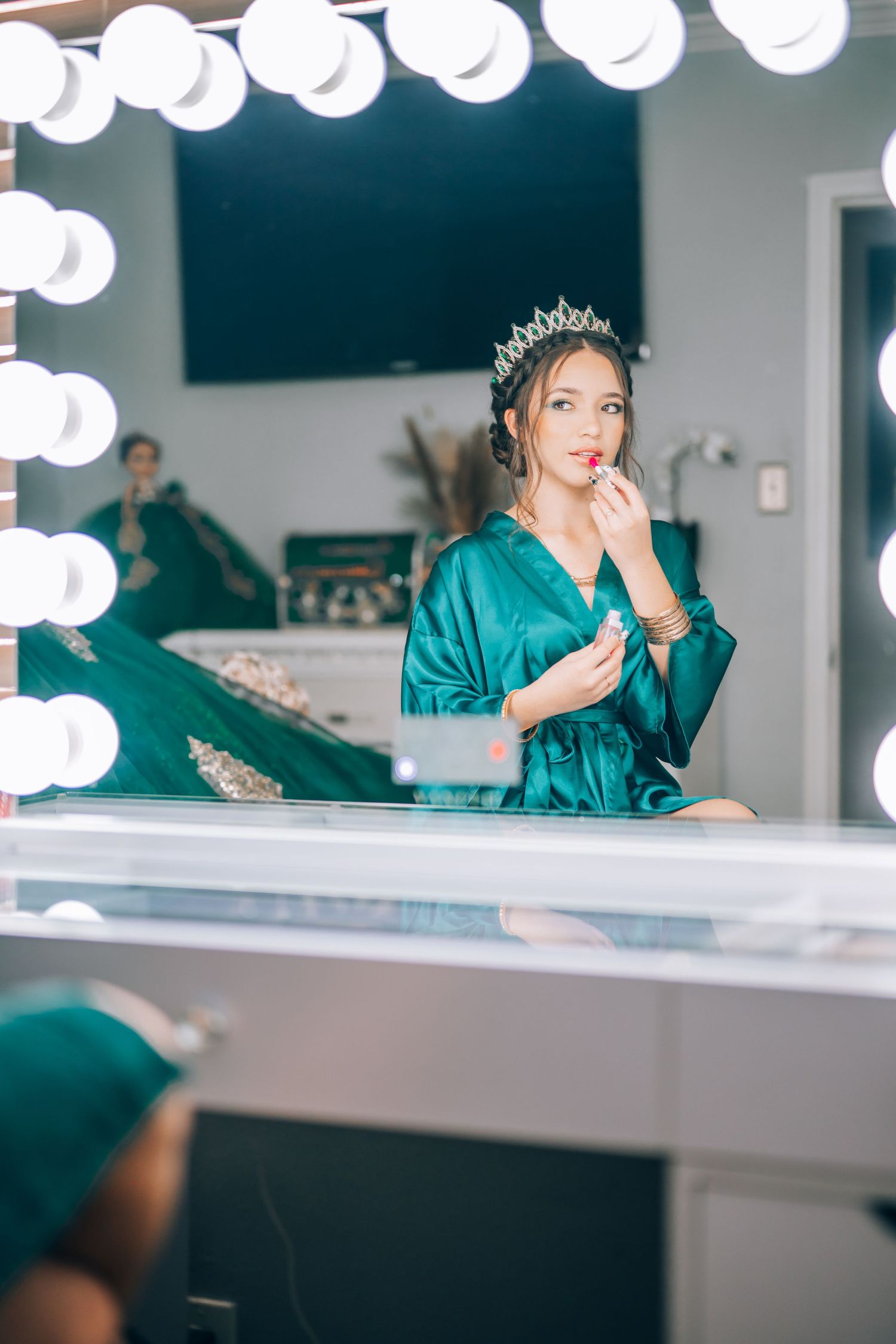Young woman wearing a crystal crown and green satin robe applies lipstick at a Hollywood vanity mirror.