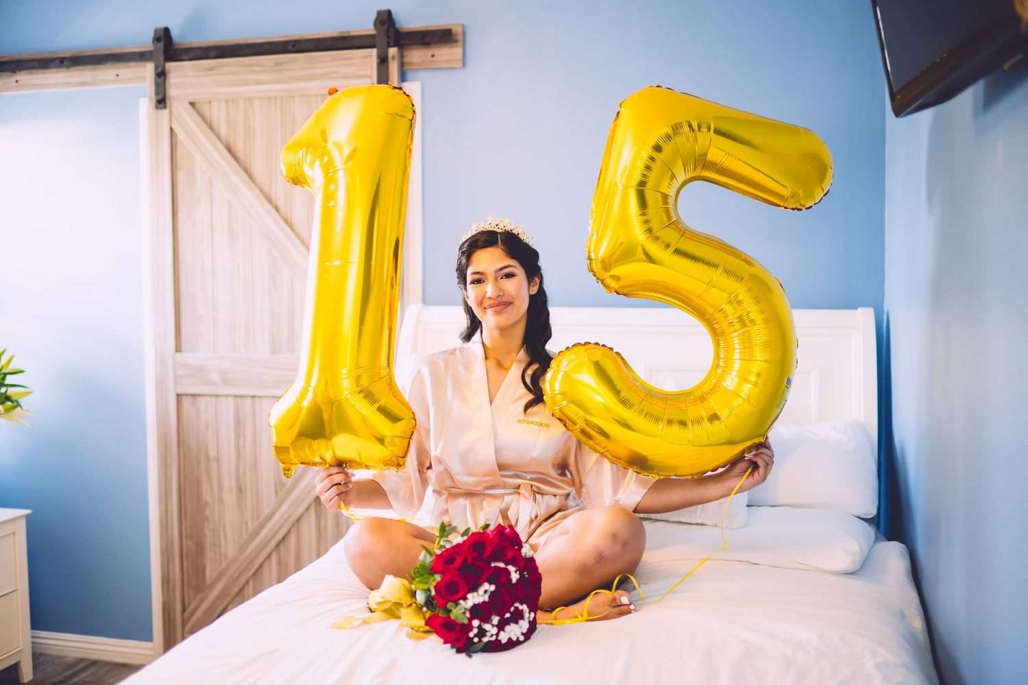 Large gold number 15 balloons displayed on a white bed with colorful floral decorations in a blue-walled room.