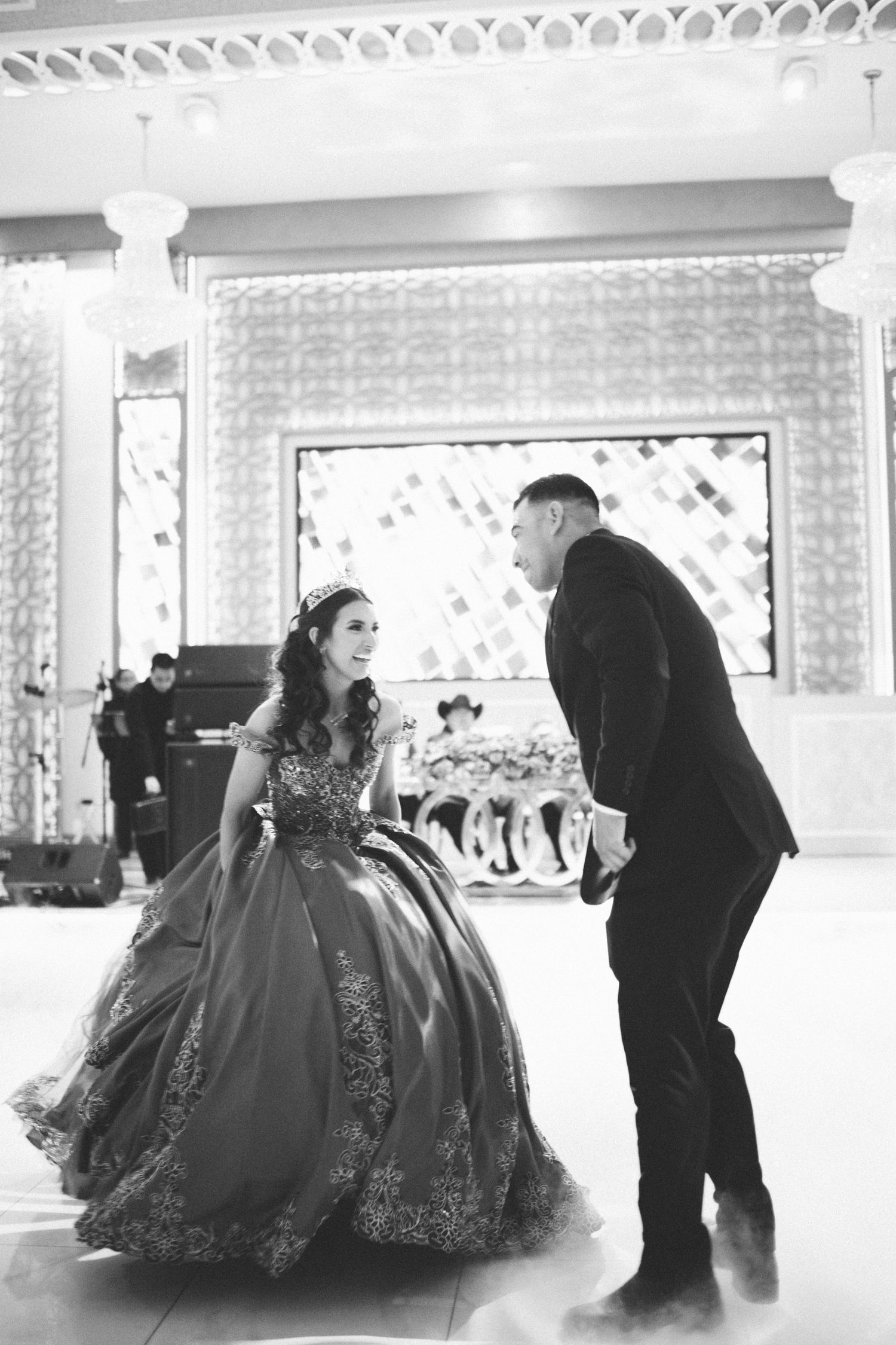 A couple in formal attire share an elegant ballroom moment, with her wearing an ornate ball gown in a grand venue.