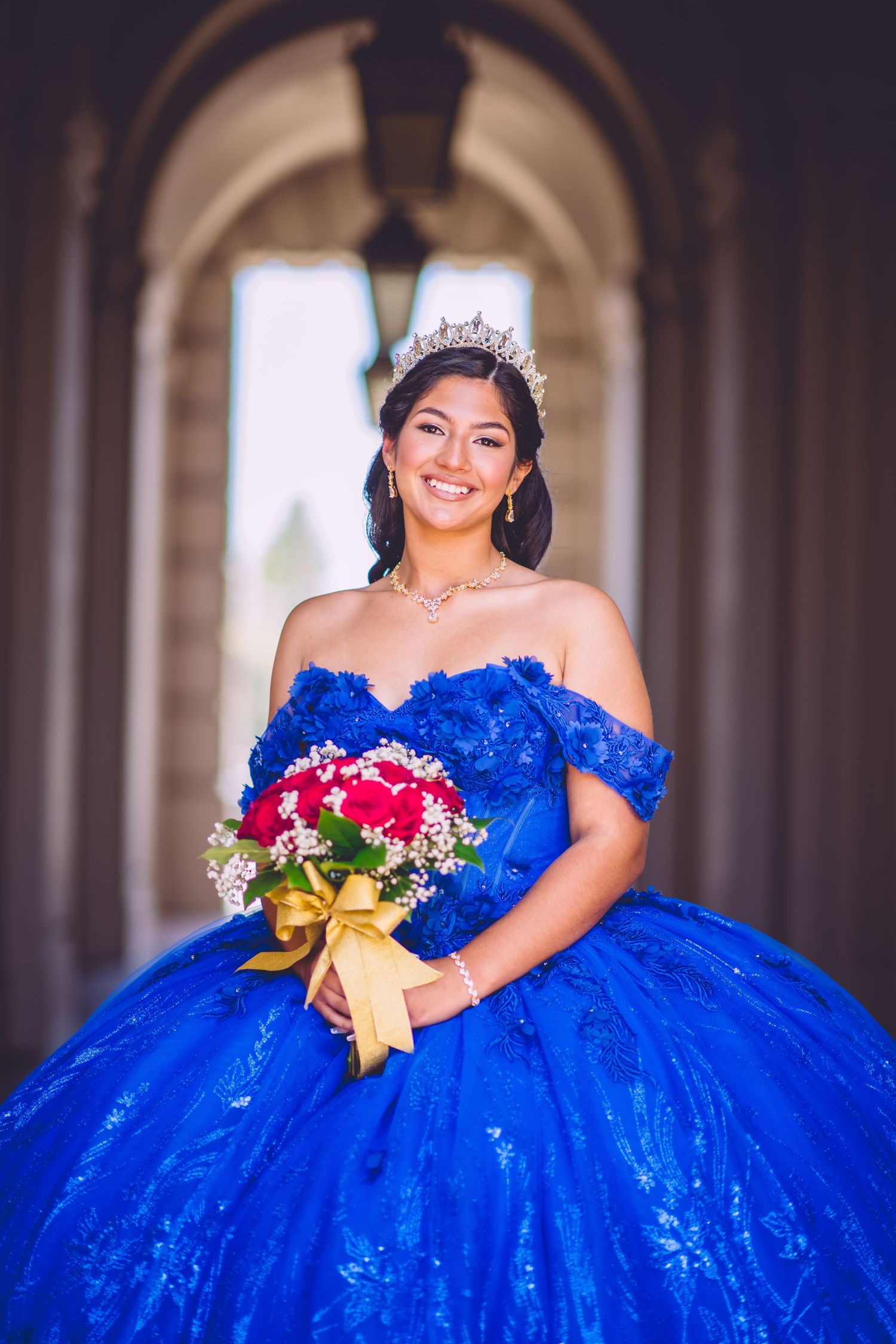 A person in a royal blue quinceanera gown with off-shoulder sleeves holds a red and white bouquet under an archway.