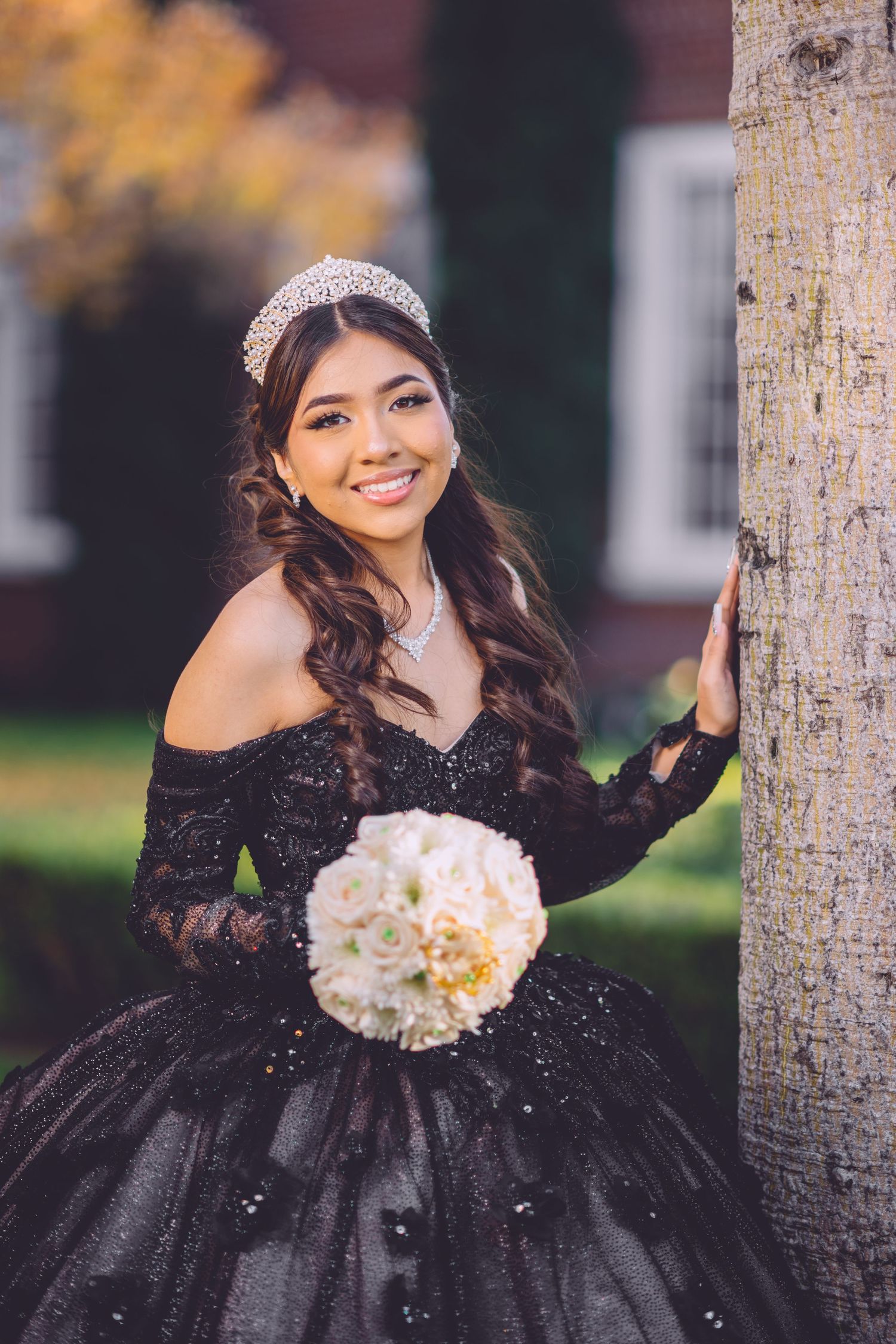 A young woman in an elegant black ball gown and tiara poses with a white bouquet next to a tree trunk outdoors.
