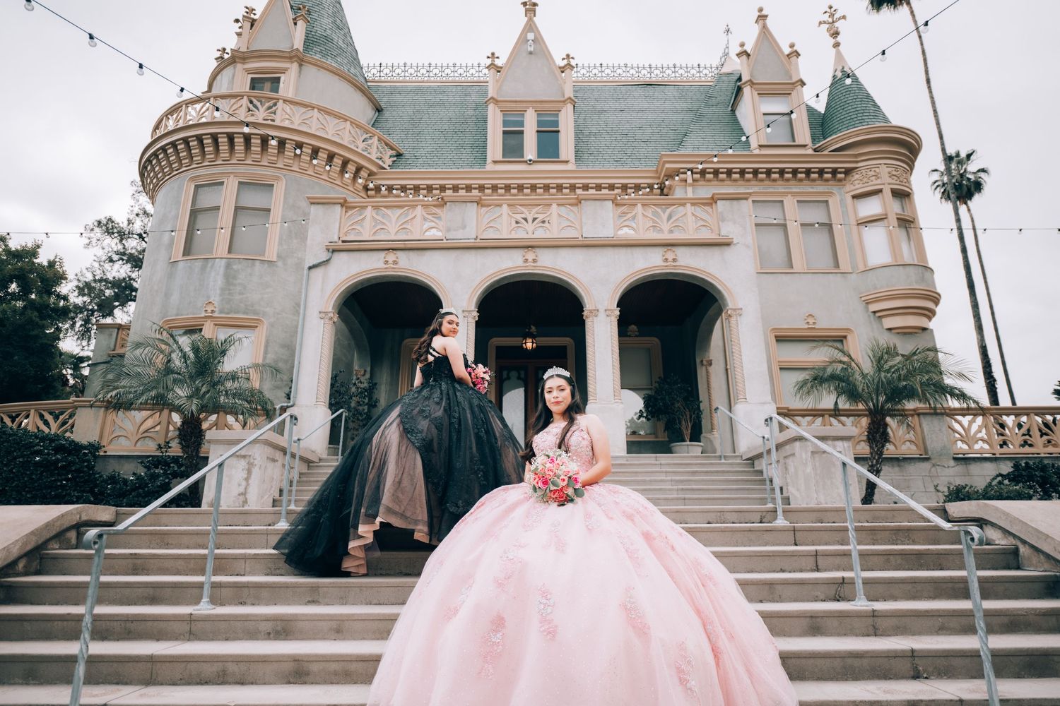 Two women in elegant ball gowns pose on castle steps, one in pink and one in black, for a quinceañera celebration.