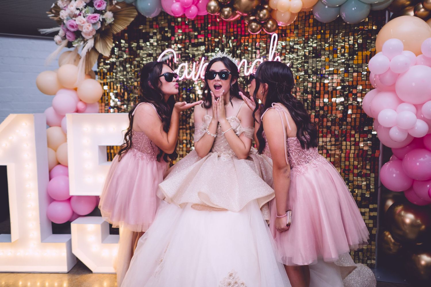 Bridesmaids in pink dresses celebrate with bride at wedding reception decorated with balloons and sparkly backdrop.