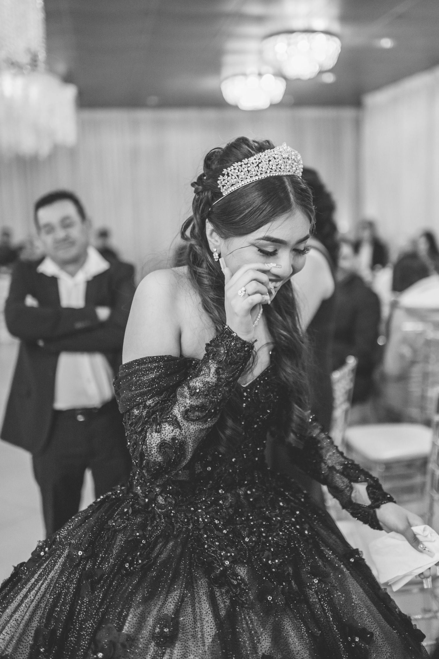 A quinceañera in a sparkly black ball gown and tiara shows emotion during her celebration as guests look on in background.