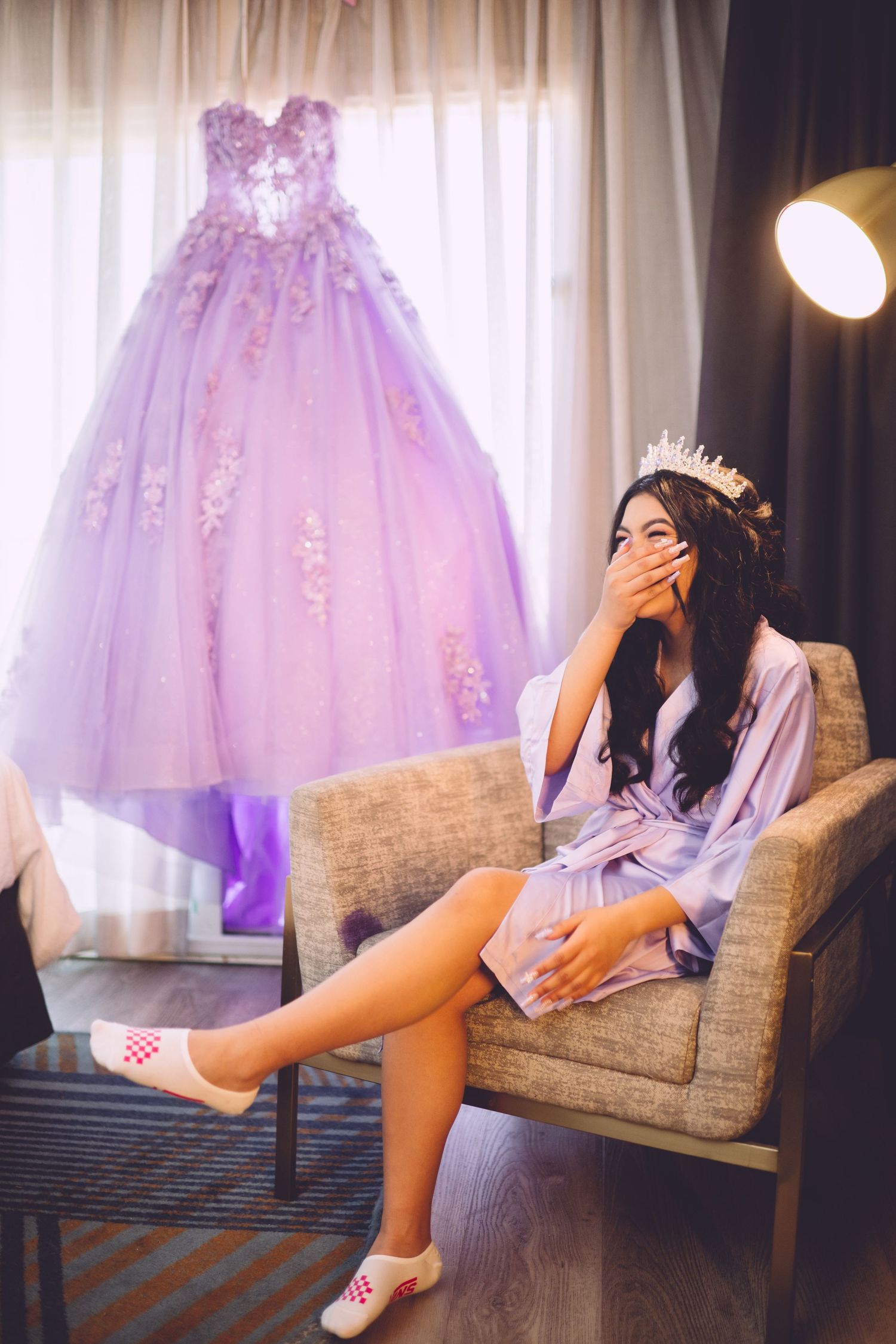 Elegant purple ball gown hangs behind couch where someone in robe and tiara sits wearing casual pink slippers.