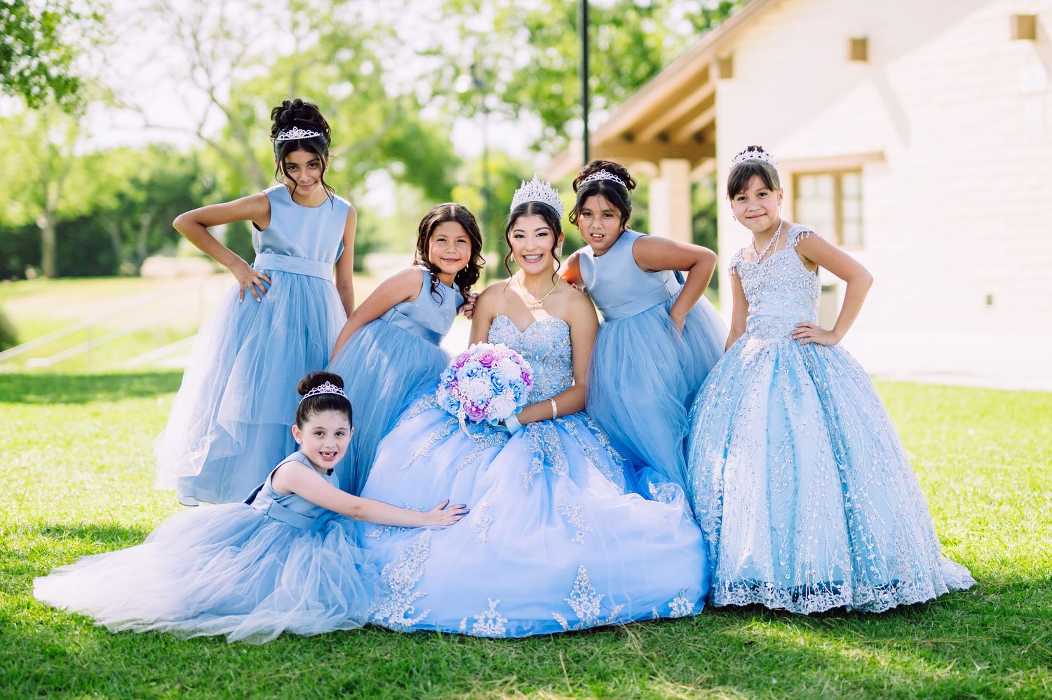 Group in flowing light blue formal dresses pose together on a sunny lawn in front of a house.