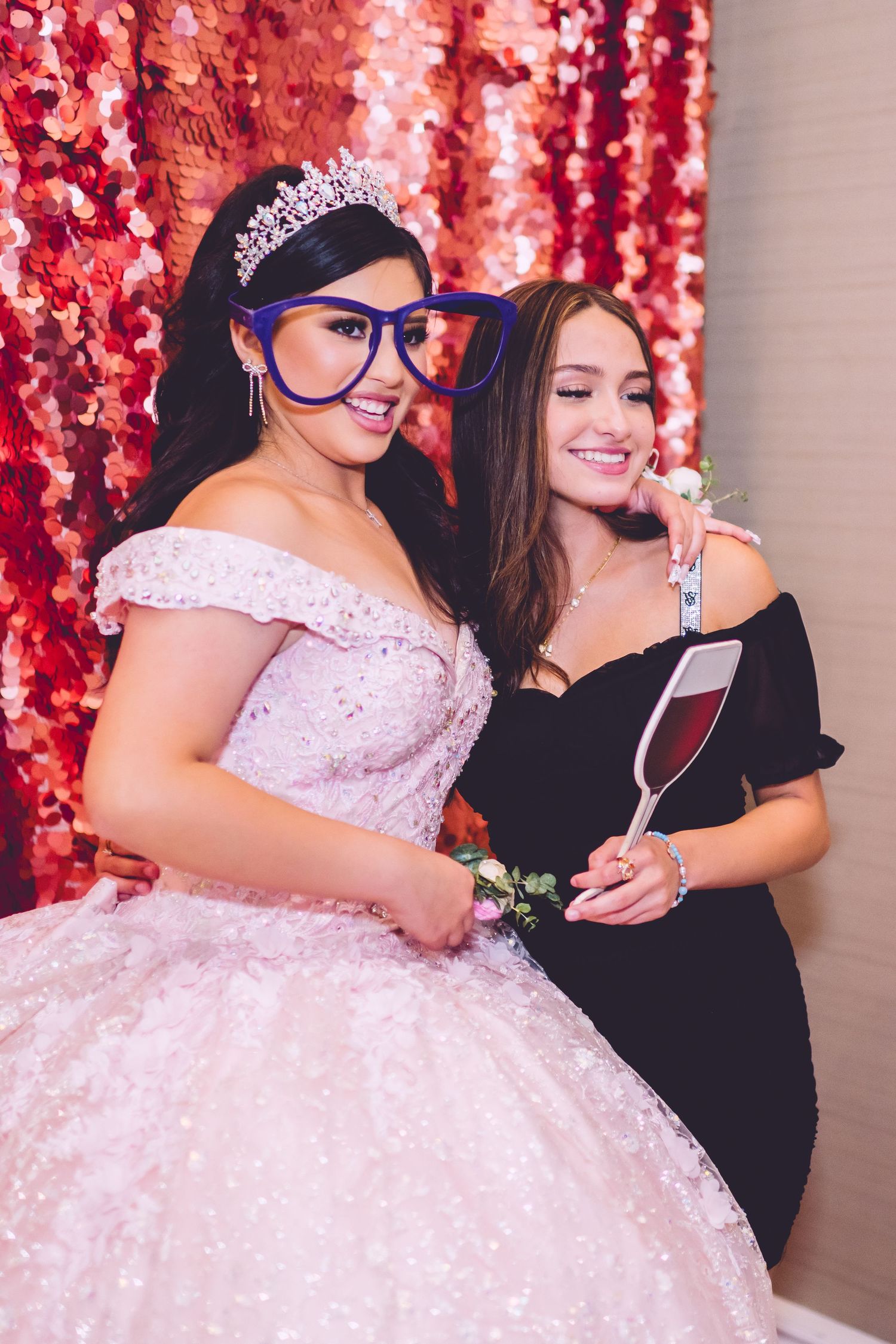 Two people at a celebration pose in front of red backdrop, one wearing pink ballgown with tiara and blue glasses.