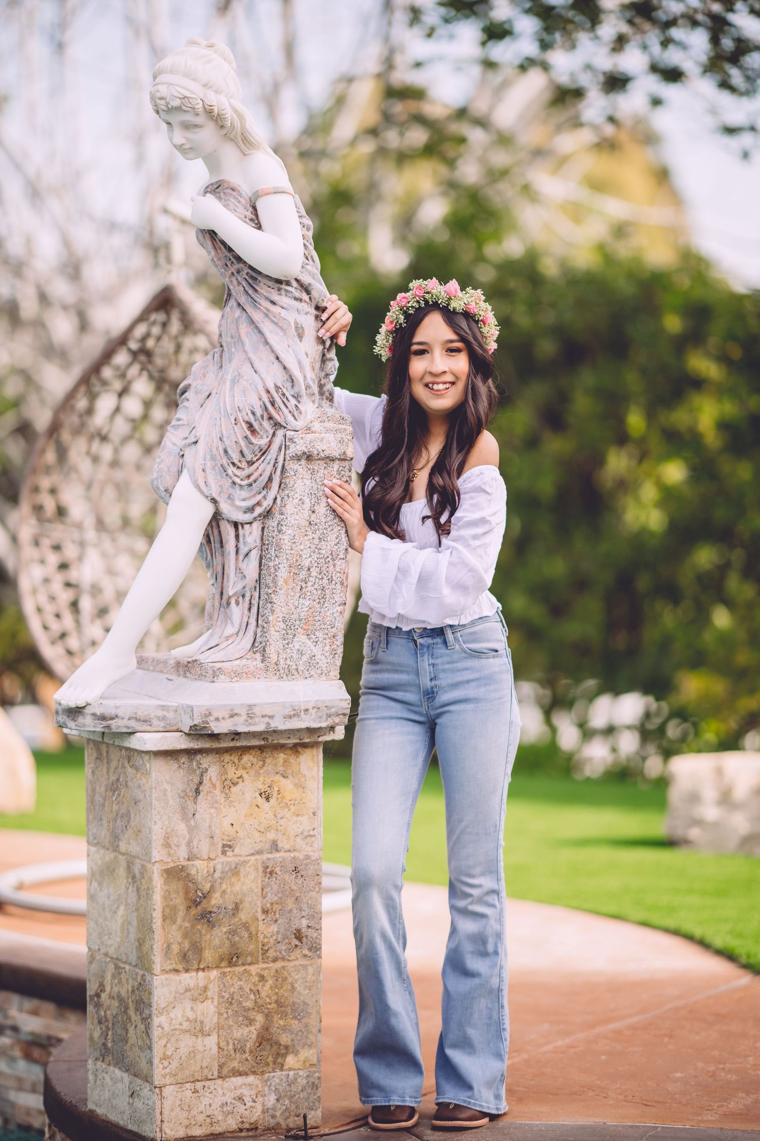 Young woman in floral crown and flare jeans poses beside classical garden statue outdoors.