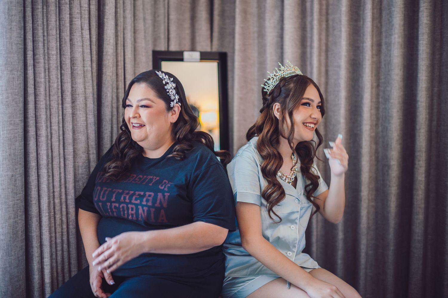 Two women sitting together, one in a Southern California tee, the other wearing a sparkling tiara, laughing in a curtained room.