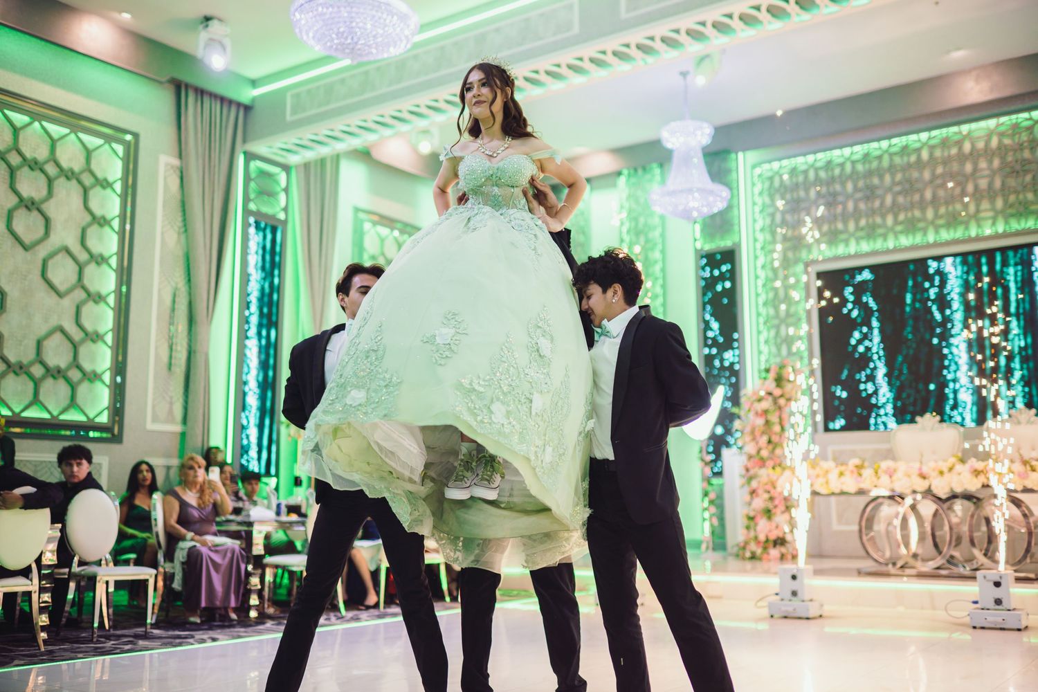 Woman in mint green ballgown lifted by two men in tuxedos on elegant decorated dance floor.