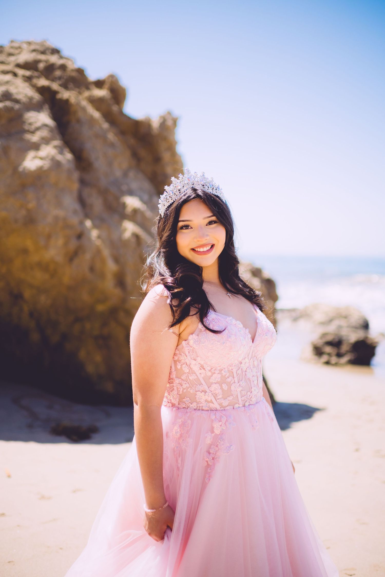 Sun-kissed beach portrait featuring a pink princess gown and sparkling crown against coastal rocks.